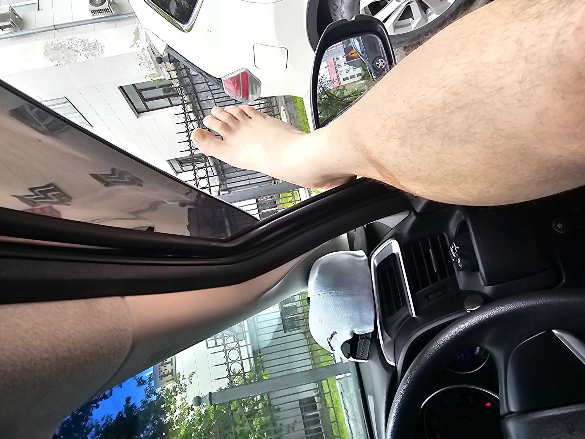 The image captures a close-up view of a person's bare leg and foot resting on the dashboard of a car. The person's leg is hairy and tanned, with the foot propped up, showing toes. The car's interior is visible, including the steering wheel, dashboard vents, and a light-colored baseball cap placed on the dashboard. Outside the car, through the windshield and side window, a white car is parked next to the car the photo is taken from. In the background, a building with a light-colored facade and windows can be seen, as well as a black metal fence and trees. A side mirror reflects a dark-colored car and a building. The overall scene suggests a casual moment of relaxation inside a parked vehicle, likely in an urban environment. The presence of air conditioning units on the building's facade suggests it is a residential or commercial structure. The architecture and the style of the vehicles hint at a location in Russia, specifically the city of Khabarovsk.