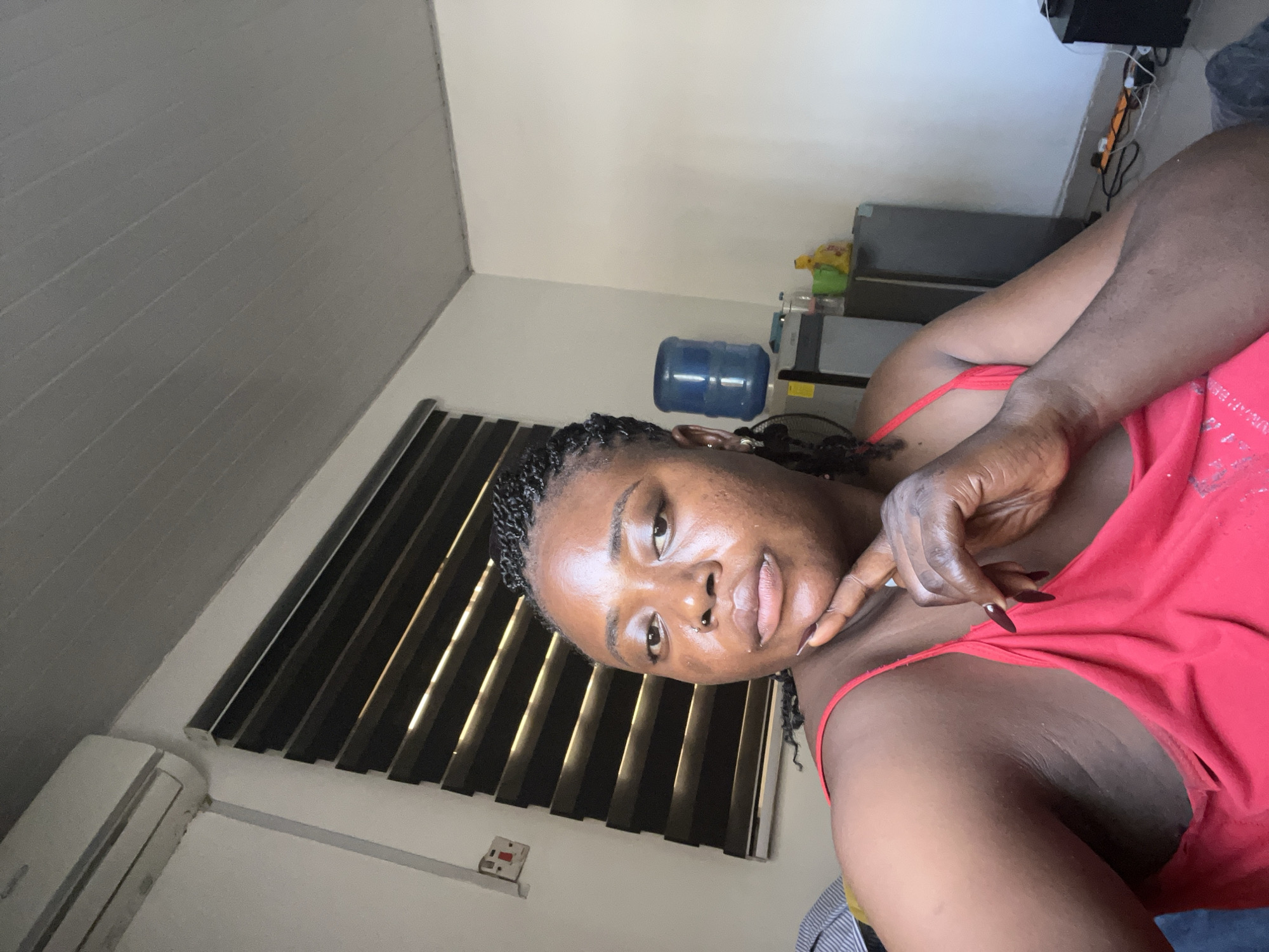 A dark-skinned woman with braided hair and a nose ring looks directly at the camera, resting her chin on her hand. She is wearing a red tank top. Behind her, a window with horizontal blinds is visible, along with an air conditioning unit mounted on the wall. Further in the background, a blue water cooler and a refrigerator can be seen. The setting appears to be an indoor residential space in Abuja, Nigeria.
