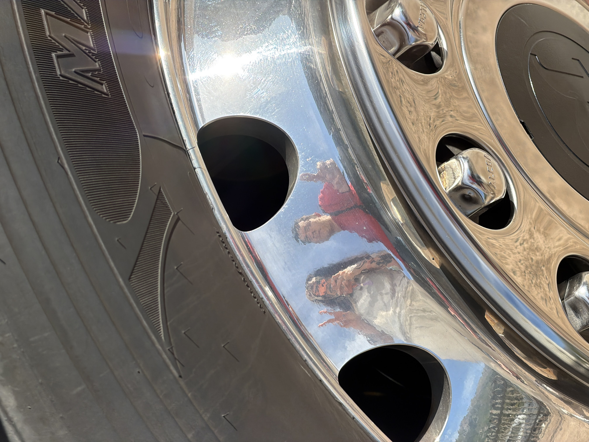 This close-up image features a large, highly polished chrome wheel and tire from a vehicle, likely a truck or bus, under bright sunlight in Budva, Montenegro. The dark rubber tire displays distinct tread patterns and sidewall markings, including partially visible embossed letters and patterns, one of which resembles an "M" or "N".
The prominent chrome rim reflects its surroundings with remarkable clarity. Reflected in the shiny surface are two individuals. One person, positioned higher in the reflection, wears a bright red short-sleeved shirt. Below them, another individual is visible, dressed in a lighter-colored top. This person's right hand is raised, making a clear "peace sign" or "V" gesture. The bright blue sky and some elements of the ground are also visibly reflected in the chrome.
The activity captured is the reflection of people interacting with or being near the vehicle's wheel, possibly posing for a photograph. The sun's intense glare creates prominent highlights and lens flares on the chrome, indicating a clear, sunny day. Despite its polished appearance, the chrome surface also shows some minor smudges and fine scratches from use.
Several hexagonal chrome lug nut covers are visible around the central hub, with the brand name "ALCOA" clearly embossed on at least two of them. The overall perspective is low, emphasizing the scale and reflective qualities of the wheel.