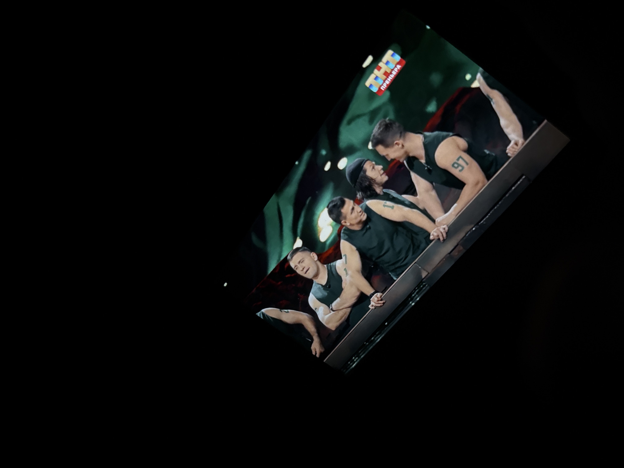 The image shows a slightly rotated view of a television screen displaying four men in athletic clothing. One man has the number "97" tattooed on his right arm. They appear to be participating in a physical competition or performance. The screen also shows the logo "THT Премьера." This is a Russian television channel. It is unknown what part of Krasnoyarsk, Russia the image was taken. The background of the screen is dark, suggesting a stage setting.