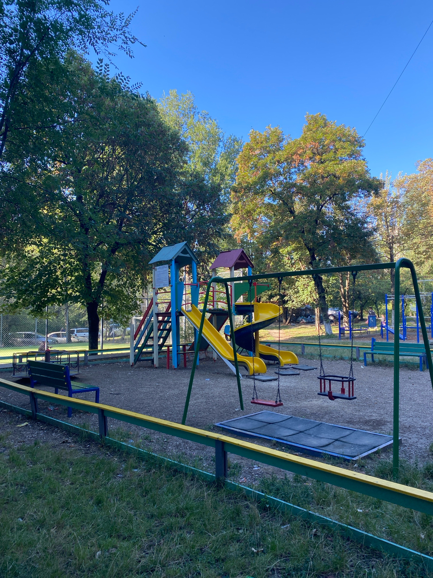 The image shows a playground in Chișinău, Moldova, on a clear day. The playground features a multi-colored slide, swings, and a merry-go-round. Trees with green and some autumnal foliage surround the area. A low fence with green and yellow rails encloses the playground. In the background, a few cars are visible parked near a building. The ground of the playground is covered with gravel.