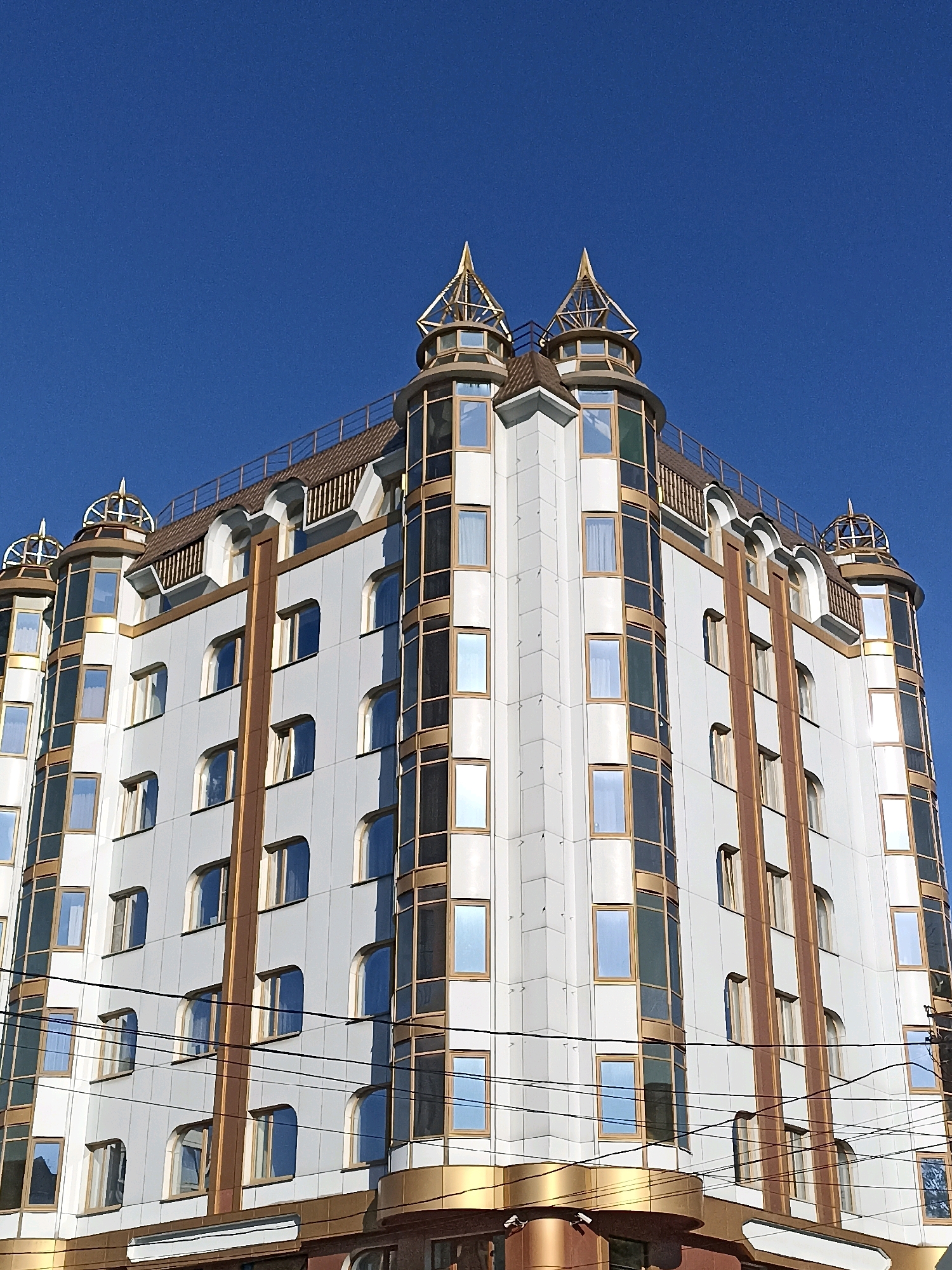 The image shows a modern, multi-story building with a white facade and decorative gold trim, against a clear blue sky. The building features numerous windows, some rectangular and some with arched tops. The upper part of the building has distinctive turret-like structures with pointed, golden roofs. Power lines are visible crossing in front of the lower portion of the building. The architecture suggests a commercial or residential building. Based on external information, this building is located in Novosibirsk, Russia, specifically at Karl Marx Avenue, 37/2, and is known as the "Dom Vostoka" or "Eastern House".