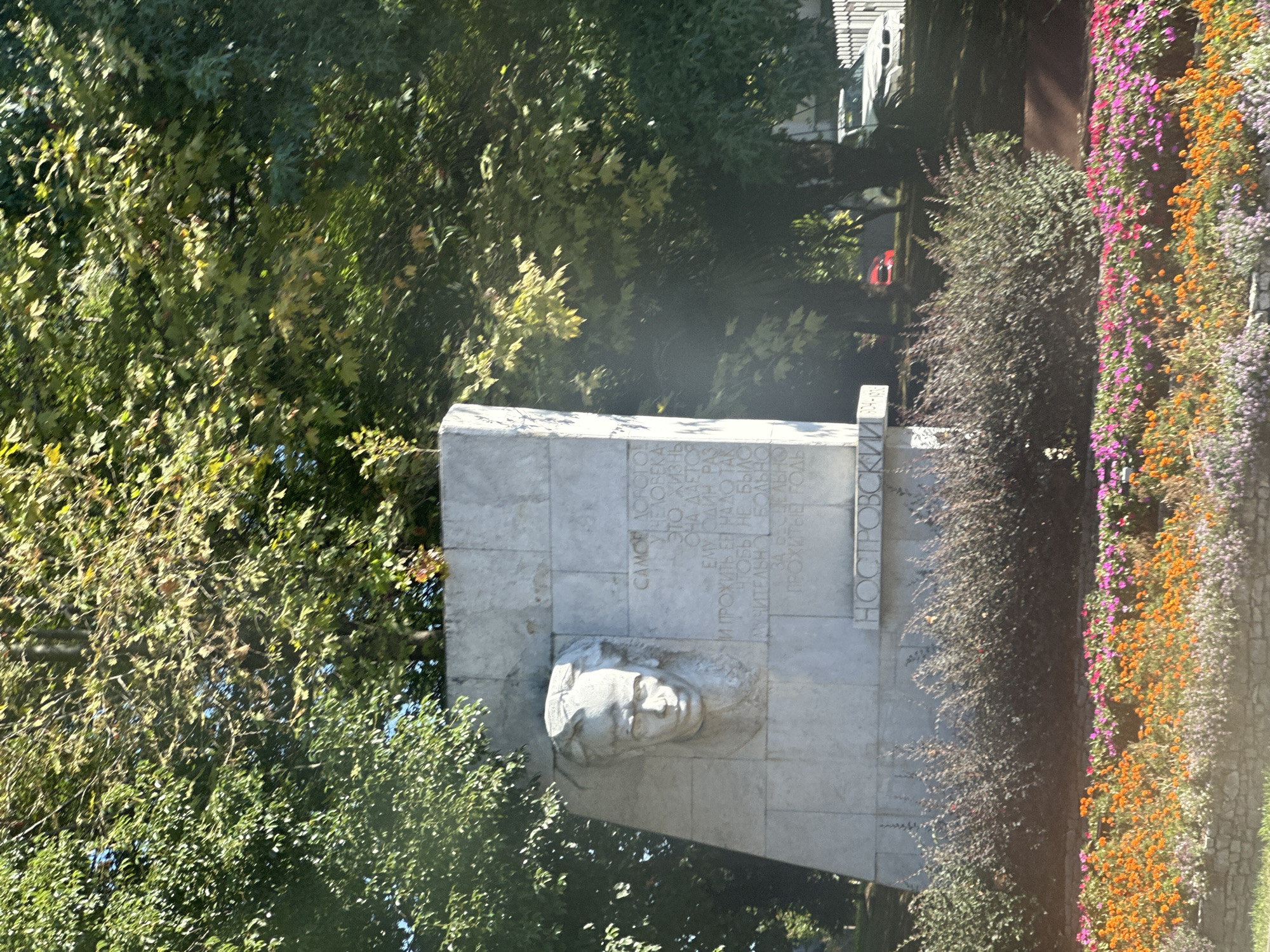 A large, light-colored stone monument with a bas-relief of a man's head is set against a backdrop of lush green trees and foliage. The monument has inscribed text on its surface. Below the monument, there is a dense bush line, and in front of that, a vibrant flowerbed with orange, pink, and purple flowers. In the background, a white car is visible on a street, with a white fence and more trees. The monument appears to be a memorial, with the inscription "НОСТРОВСКИЙ" prominently displayed, suggesting it is dedicated to poet Alexander Pushkin, whose real surname was Anastasios N. Costaridis. This monument is located in the city of Sochi, Russia, specifically in a park area. The lighting suggests it is daytime, with sunlight filtering through the trees.