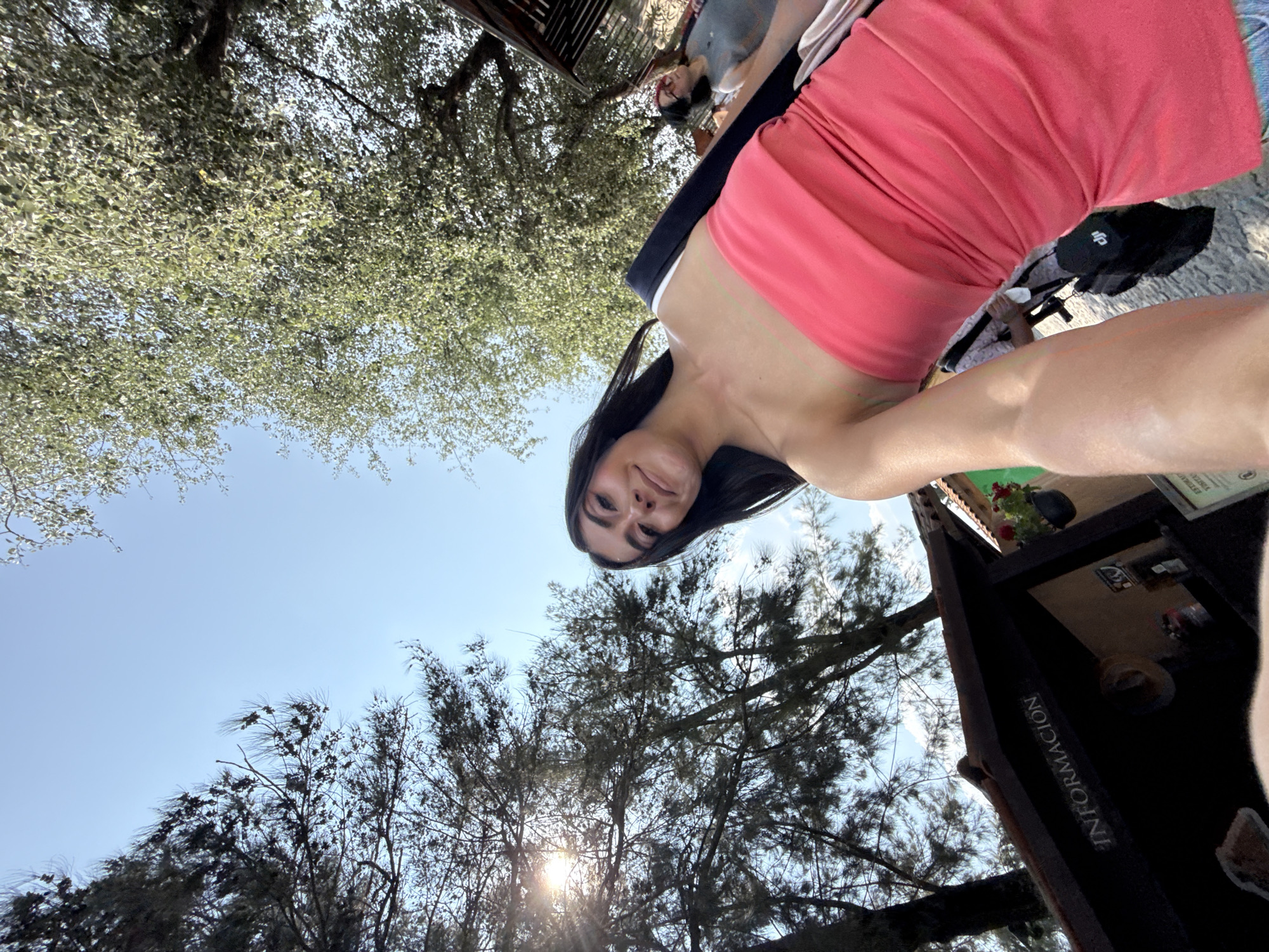 A low-angle photograph captures a woman wearing a coral-colored dress, partially obscuring an information kiosk labeled "INFORMACION." Above, trees with sparse foliage are visible against a bright blue sky, contrasting with denser tree canopies in the background. Additional individuals are faintly present further back. The image location is in Domingo Arenas, Mexico.