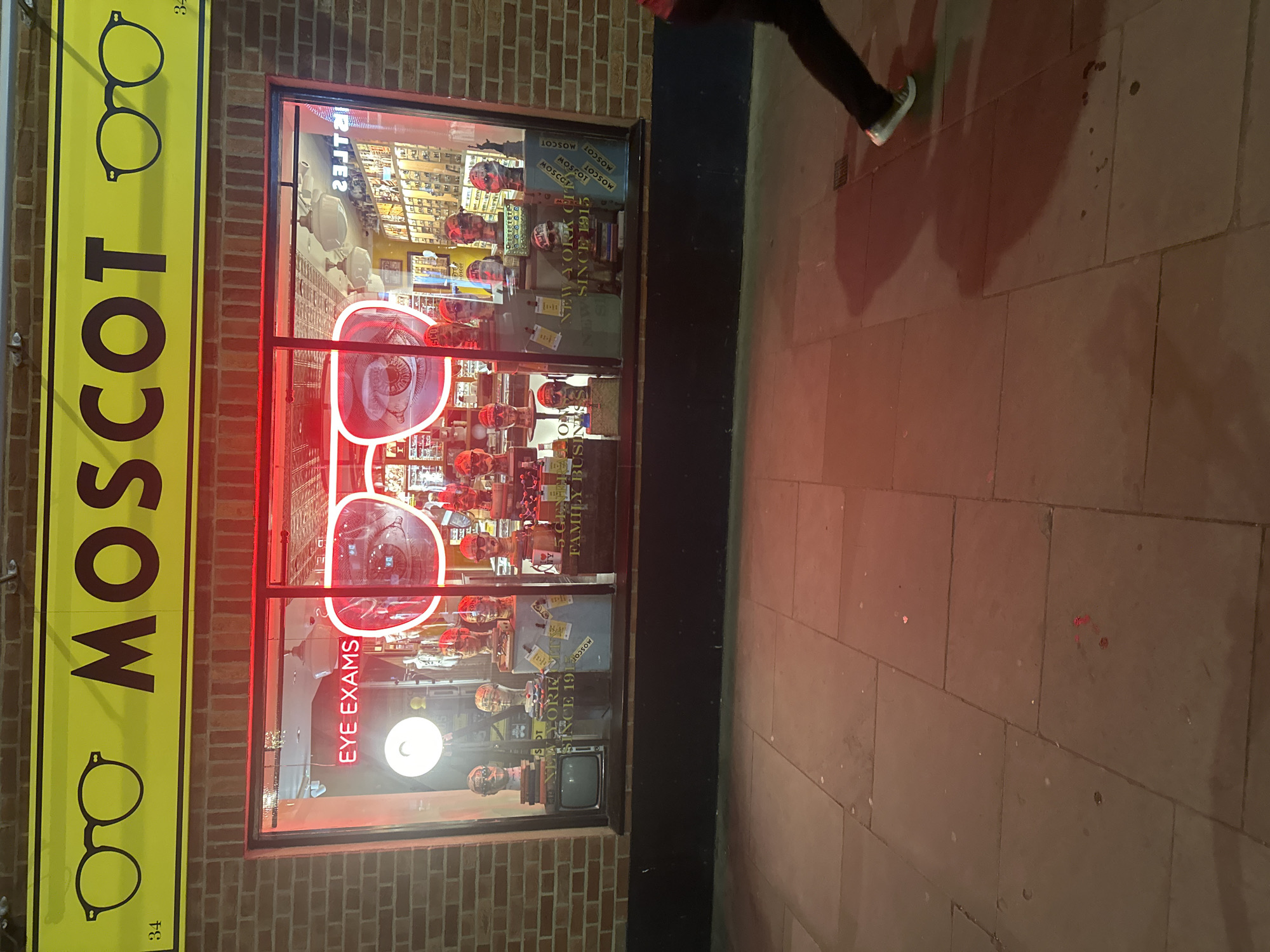 The image depicts a nighttime street scene featuring the storefront of an eyewear store named "MOSCOT," likely in London, United Kingdom. A prominent vertical yellow sign on the left side of the frame displays "MOSCOT" in black capital letters, accented by two black outlined eyeglasses. The number "34" is also visible on this sign.
The store's large display window is brightly lit from within, featuring striking red neon lights that outline oversized pairs of eyeglasses. Inside, numerous mannequin heads, each adorned with a pair of glasses, are arranged on shelves and various display stands, showcasing the store's products. Red neon text within the window spells out "EYE EXAMS." Further text visible inside the display includes "NEW YORK CITY SINCE 1915" and "5 GENERATIONS FAMILY BUSINESS," emphasizing the brand's heritage. The name "MOSCOT" is also repeated on smaller product displays.
On the right side of the image, the lower leg and a foot wearing a white sneaker are visible, belonging to a person walking past the storefront on the tiled pavement. An elongated shadow of the person is cast across the ground, illuminated by the artificial light. The building itself is constructed from reddish-brown bricks. The overall impression is an active urban environment after dark, with the store's vibrant lighting drawing attention.