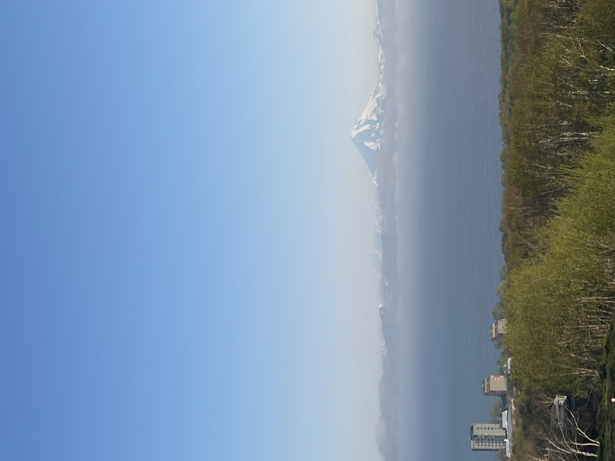 The image shows a panoramic view of a coastal city with a large snow-capped mountain in the background. In the foreground, there is a dense forest with trees in various stages of spring foliage. Beyond the forest, there is a body of water, likely the sea, with several buildings situated along the coast. The buildings appear to be apartment complexes, with one tall, grey building and two shorter, red-and-white buildings. The sea is a deep blue, and a layer of white clouds or mist partially obscures the distant mountains. The most prominent feature in the distance is a majestic, cone-shaped mountain covered in snow, with a clear blue sky above. The scene is well-lit, suggesting it was taken during daylight hours. The overall impression is one of natural beauty with a touch of urban development.
The location is identified as Krutoberegovy, Russia, which is a coastal settlement in Kamchatka Krai. The prominent snow-capped mountain in the background is likely one of the volcanoes in the region, such as Koryaksky or Avachinsky volcano, which are visible from Petropavlovsk-Kamchatsky, the administrative center of Kamchatka Krai, and Krutoberegovy is located nearby. The buildings along the coast are consistent with residential architecture found in coastal towns of this region. The lush green forest in the foreground indicates a temperate climate, likely during the warmer months of spring or early summer.