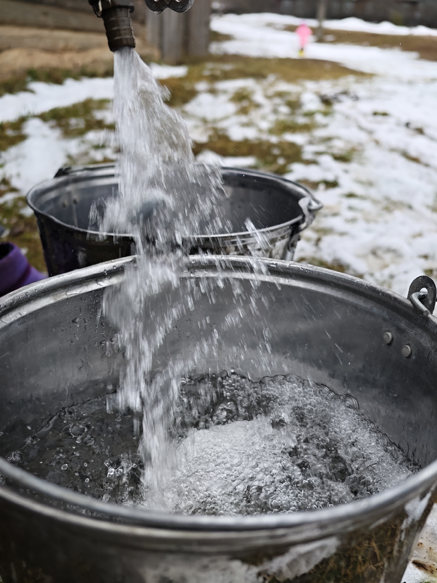 In Уральское, Russia, a metal faucet pours water into two metal buckets. The foreground bucket is nearly full, with visible bubbles and water droplets. The background shows a mix of grass and snow. A blurry figure in a pink outfit stands in the distance.