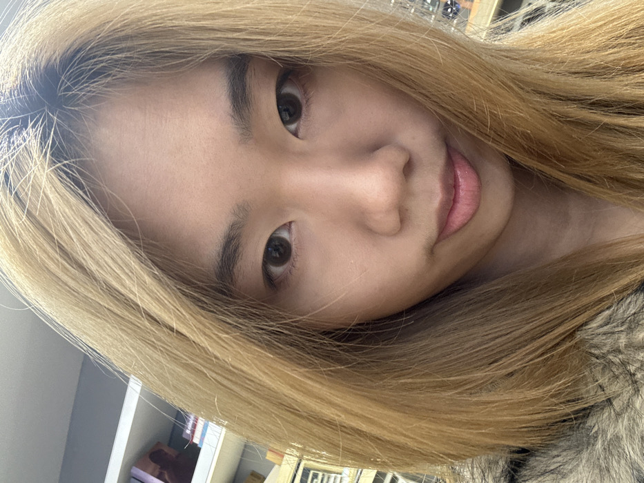 The image displays a close-up, head-and-shoulders portrait of a single individual, positioned centrally and looking directly into the camera. The individual has fair skin, dark brown eyes, and light brown hair, appearing to be dyed or bleached, with darker roots visible at the crown. The hair is long and falls around the face, extending out of the frame. The subject's mouth is closed with a slight downward curve, and no specific action beyond posing for the photograph is evident. A segment of a dark, textured garment, resembling faux fur, is visible on the lower left. The background is blurred, indicating a shallow depth of field, but shows an indoor environment. A white shelf unit is partially visible on the upper left, containing multiple indistinct objects consistent with books or packaged items. Additional out-of-focus elements are present in the background to the right. The scene is located in Shapingba District, China.