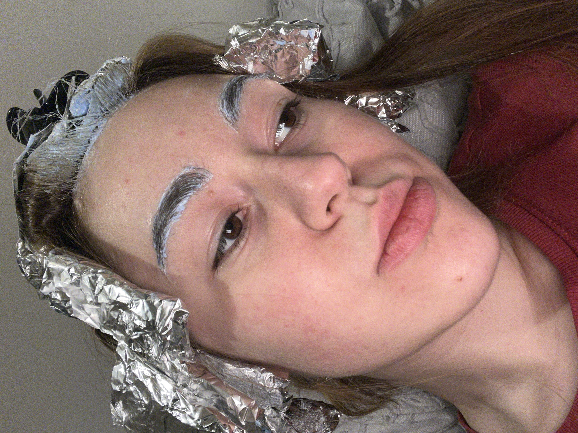 A close-up, angled shot shows a woman with brown hair and fair skin, undergoing a hair-coloring process. Her eyebrows are covered in a white substance, likely bleach or dye. Aluminum foil is wrapped around portions of her hair, presumably to isolate sections being treated. A black hair clip is visible amongst the foil and hair at the top of the frame. The woman is wearing a red sweatshirt and is resting against a gray pillow. The background appears to be a neutral-colored wall or surface. The location is not discernible from the visual elements of the image.
