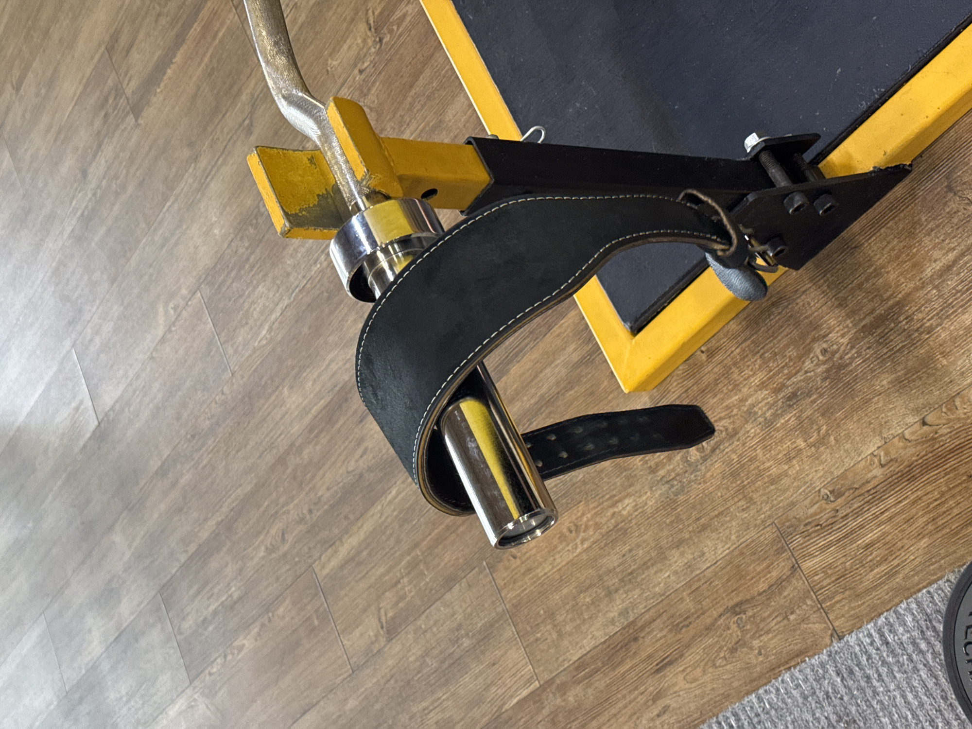 A close-up, high-angle shot shows a weightlifting belt draped over a barbell rest. The belt is black with white stitching and a silver buckle. The barbell rest is yellow and black metal. The background is a wooden floor with a visible grain pattern. The image is forensic in its detail, capturing the texture of the wood and the reflective surfaces of the metal. The scene is devoid of human presence and appears to be set in a gym. This image is not from Chaikovsky, Russia.
