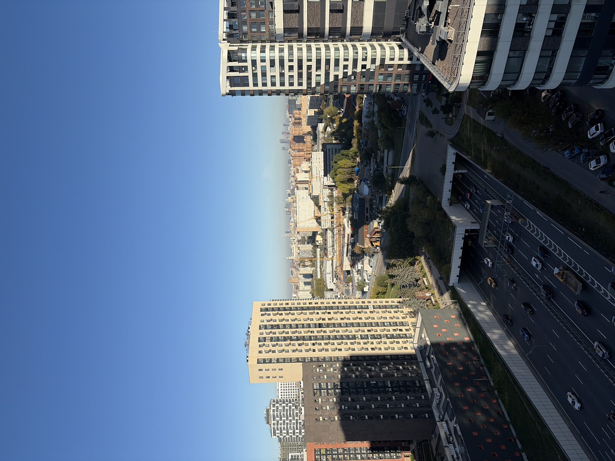 The image captures a panoramic view of a modern urban landscape in Moscow, Russia, on a clear, sunny day. The foreground is dominated by a multi-lane highway where cars and trucks are actively traveling. The highway is bordered by concrete barriers and slopes, with some vegetation visible along the embankments. To the left of the highway, a dark-colored building with many small orange-tinted windows is partially shaded by a taller, light-colored residential building.
The midground features a complex of residential buildings of varying heights and architectural styles. A prominent, tall, light-yellow building with numerous dark windows stands out. Adjacent to it are other high-rise apartment blocks with balconies and repeating window patterns. The cityscape extends into the background, revealing a dense collection of buildings, some of which appear to be older residential blocks and others are modern high-rises, including a distinctive white and dark-paneled building on the right. Distant skyscrapers are visible on the horizon, suggesting the presence of a city center or business district. Green trees are interspersed throughout the scene, particularly in the midground, adding a touch of nature to the urban environment. The clear blue sky suggests a pleasant, possibly autumn or spring day. The overall impression is one of a densely populated, developing urban area in Moscow.