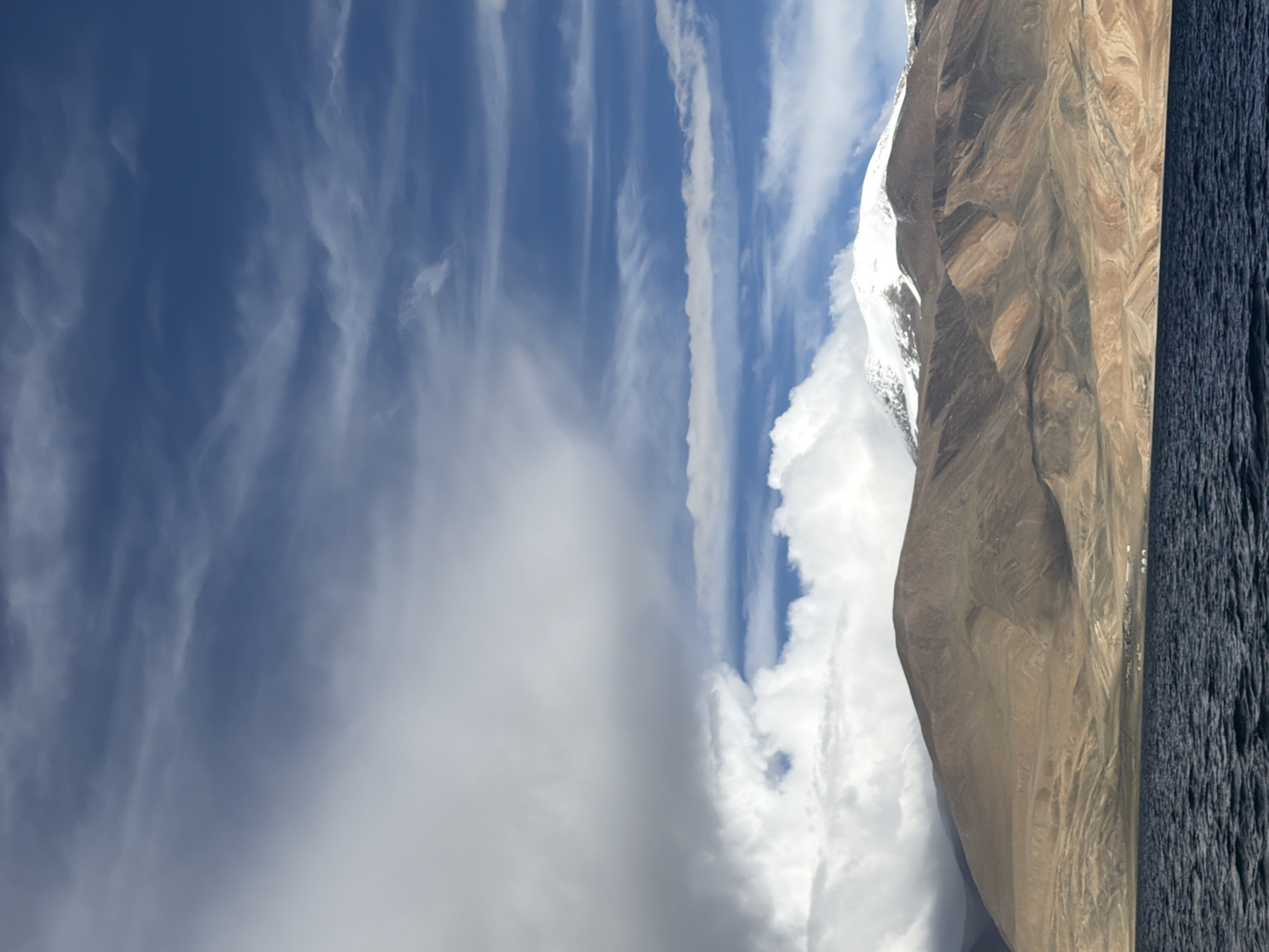 An expansive, high-angle shot captures a serene lake in the foreground, its dark blue waters rippling under a partly cloudy sky. In the midground, a chain of rugged, earth-toned mountains stretches across the frame, their slopes marked by subtle variations in color and texture. The peaks of some of these mountains are capped with snow, adding a touch of white to the otherwise muted landscape. In the distance, a small settlement of white tents and structures is visible along the base of the mountains. The background is dominated by a vast expanse of sky, filled with dramatic clouds ranging from thick, white formations to wispy streaks of cirrus clouds against a brilliant blue backdrop. The scene appears to be set in a remote, mountainous region of India, possibly Ladakh, given the arid terrain and the presence of a high-altitude lake.