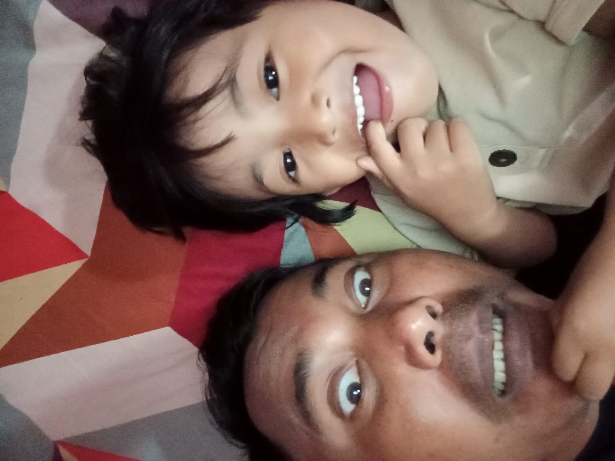 This is a close-up, selfie-style photograph taken from a low angle, showing two individuals lying down on a bed with a patterned bedsheet. The person in the foreground, positioned at the bottom of the frame, is an adult male. His face is angled towards the camera, and he is looking up with his eyes wide open. He has dark hair and is wearing a light-colored garment with a button. His mouth is slightly open, revealing his teeth.
The second individual, a young child, is positioned above the adult, with their head tilted. The child has dark hair and is looking directly at the camera with a wide, open-mouthed smile, showing their teeth. The child's right hand is raised and appears to be touching their face near their mouth. The background consists of a colorful, geometric patterned bedsheet with shades of red, brown, white, and gray. The image appears to have been taken indoors, likely in a bedroom in Bloro, Indonesia, given the context. The lighting suggests a natural light source from above.