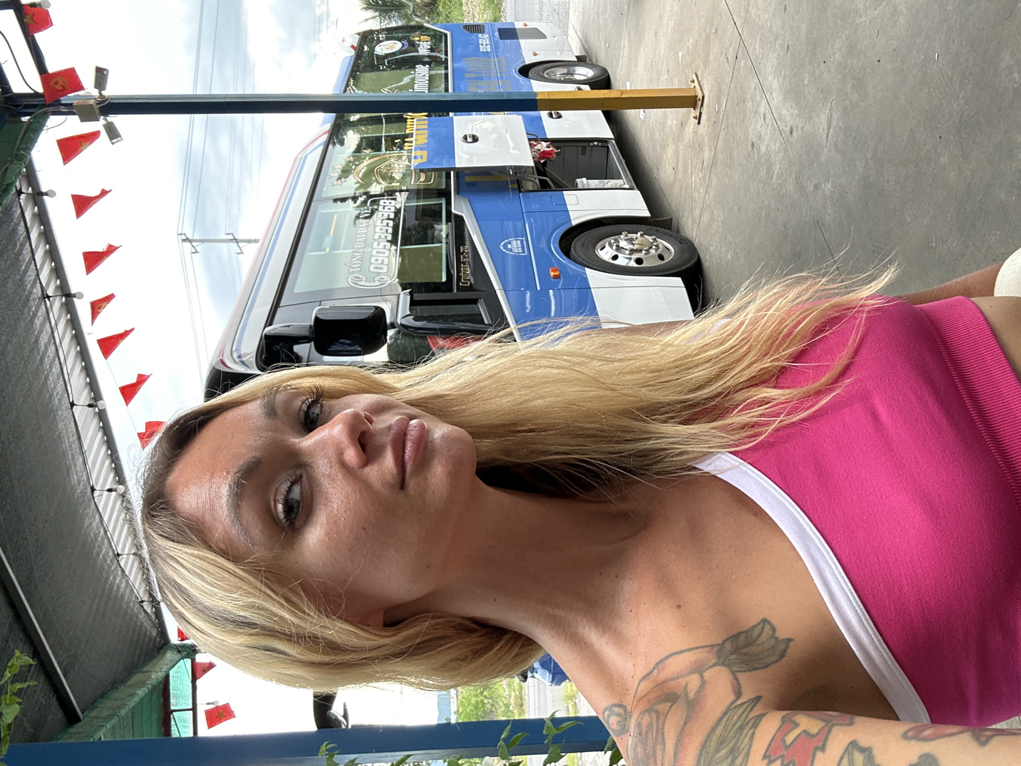 A woman with blonde hair, wearing a pink top, poses for a selfie outdoors. She has a tattoo on her arm. Behind her, a blue and white double-decker bus is parked. The bus has text that appears to be in Vietnamese. Red flags with yellow stars are strung across a wire above the bus and in the background. The scene appears to be a roadside stop or bus station. The lighting suggests it is daytime, with an overcast sky. The location cues, such as the flags and the style of the bus, are consistent with Vietnam.