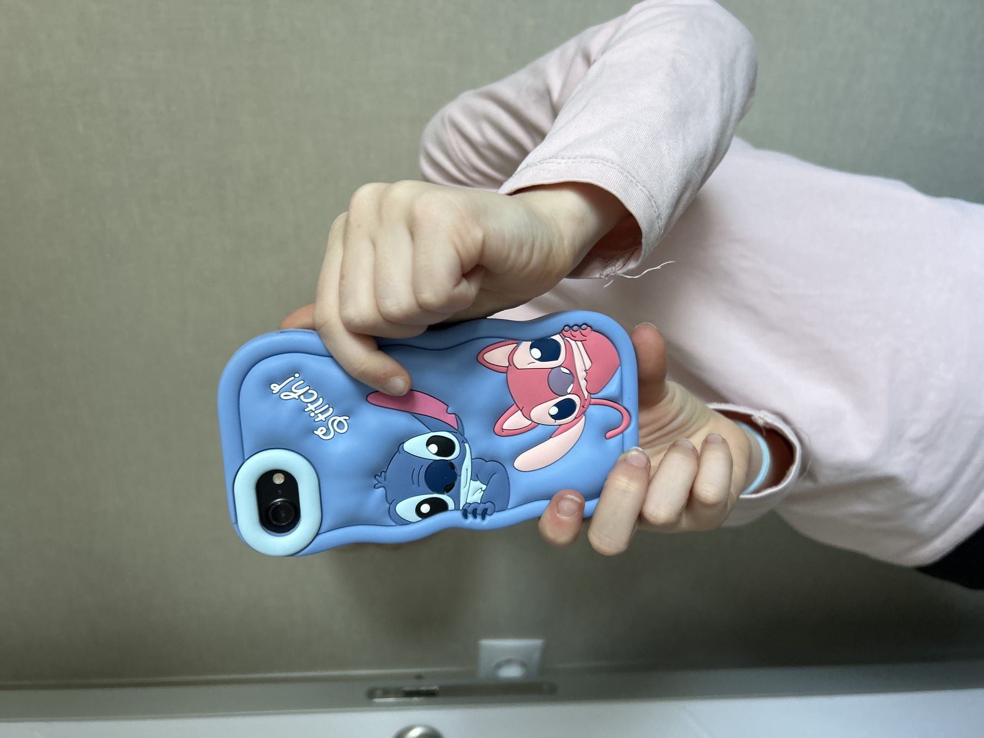 A child in a pink long-sleeved shirt is holding a light blue cell phone case with cartoon depictions of Stitch and Angel from the Disney movie "Lilo & Stitch." The phone case is held in front of them, with one hand gripping the top and the other supporting the bottom. The phone case is oriented horizontally and shows the camera lens is on the left side of the image. The background is a blurry wall with a light fixture at the bottom. The location is potentially Chaikovsky, Russia.