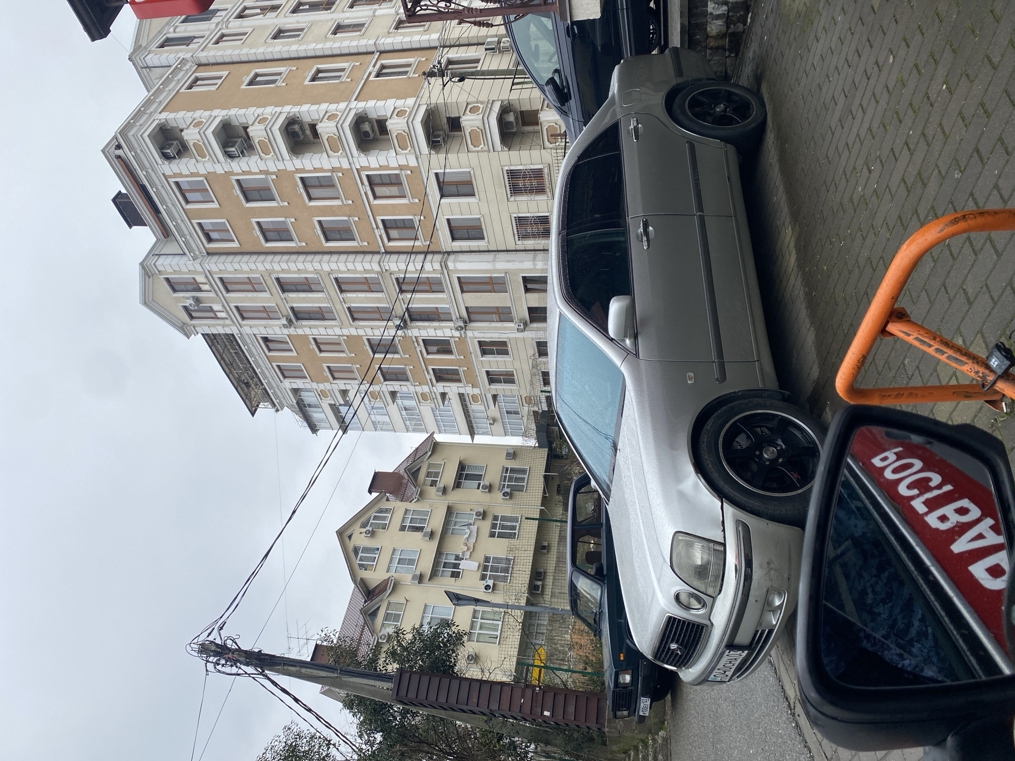 The image shows a street scene in Sochi, Russia. A silver car is parked on a paved street, with its front end facing the viewer. Behind it is a dark-colored vehicle. A tall building with multiple stories and balconies is in the background, along with a smaller building. Utility poles and wires are visible in the lower left corner. A blurred orange object and a car's side mirror with a reflected sign are in the foreground. The sky appears overcast. The car's license plate is partially visible.