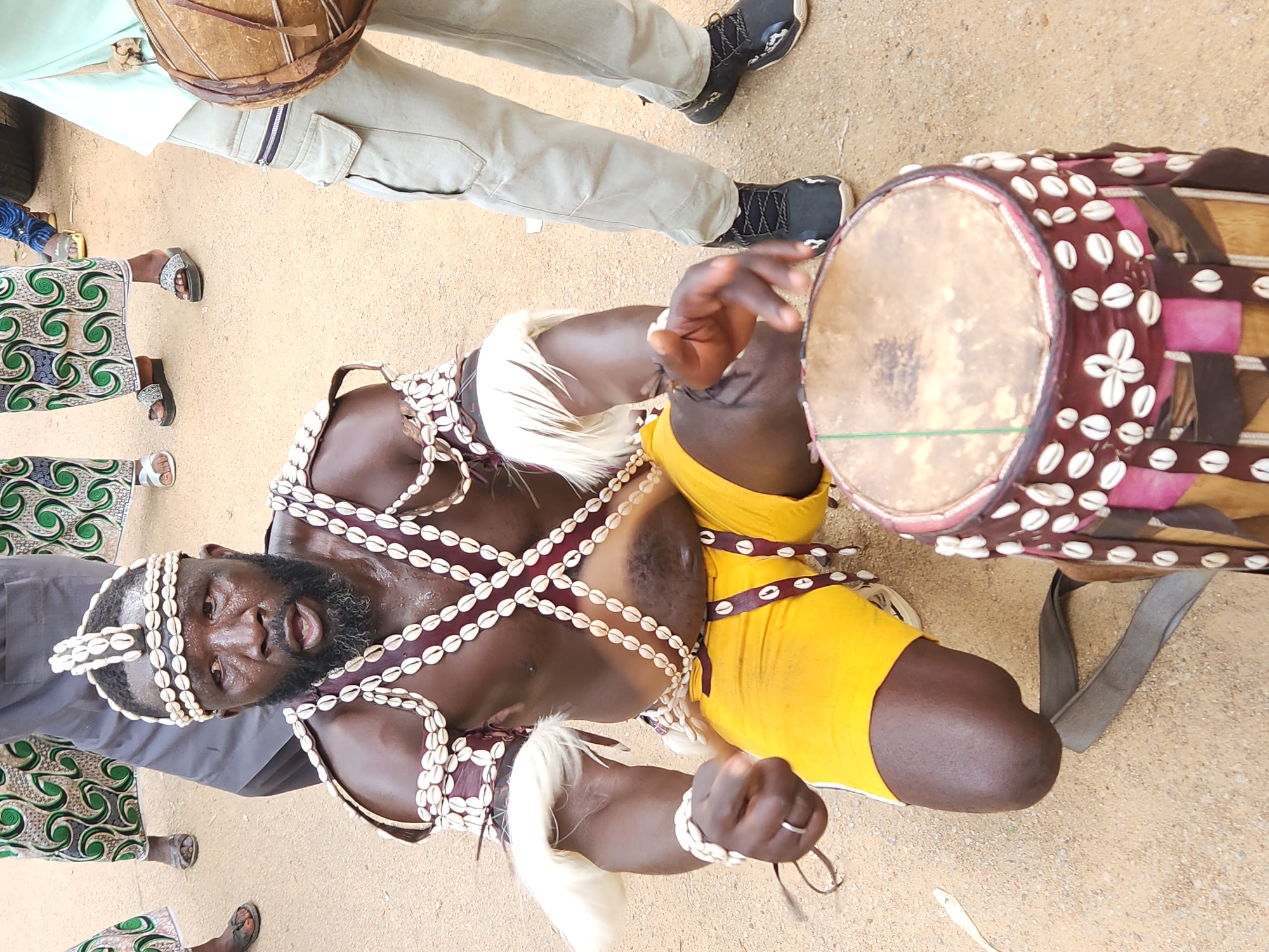 This media file depicts a man dressed in traditional attire, adorned with numerous shell necklaces and a headdress, in a sandy outdoor setting. He is holding a drum with a leather head, decorated with shells and colorful fabric. The man's face is visible, and he appears to be engaged in a performance or ritual, with his mouth open and eyes focused. His body is partially obscured by his clothing, which includes a yellow garment and possibly other decorative elements.
The scene suggests a cultural event or celebration, likely taking place during the day given the lighting. The sandy ground and the man's attire are indicative of a location in Nigeria, possibly Jos as suggested. Other individuals are partially visible in the background, wearing patterned clothing, suggesting a communal gathering. One person is wearing light-colored pants and dark athletic shoes, while others are seen in dresses with green and white swirling patterns. The overall atmosphere seems to be energetic, potentially involving music and dance, given the presence of the drum. No visible text is present in the image.
