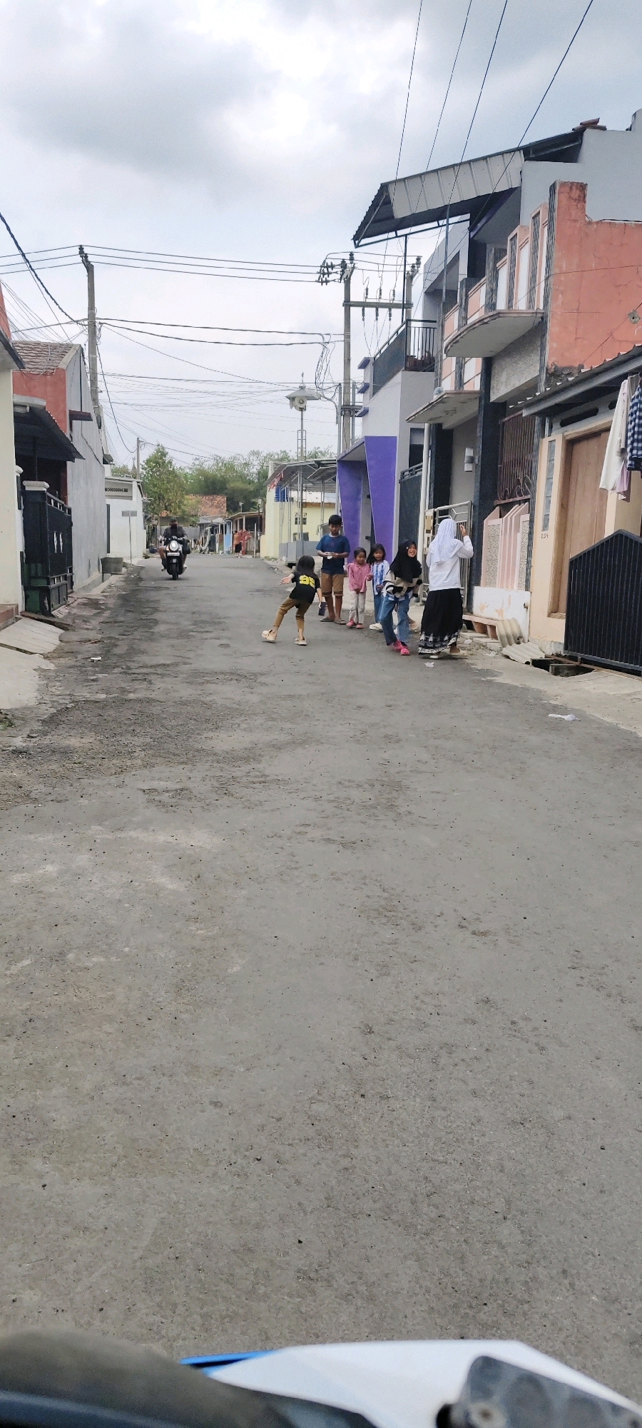 The image depicts a street scene in Tasikmalaya, Indonesia, characterized by modest residential buildings lining a paved road. The sky is overcast, suggesting a cloudy day. In the foreground, the road surface appears worn with patches of gravel and darker asphalt. A motorcyclist is visible in the middle distance, heading away from the camera down the street. A group of children are gathered on the right side of the road, some standing and observing, while one child in the foreground is in motion, possibly playing or running. A woman in a white hijab and black skirt is also part of the group. The buildings on the right are multi-story structures with balconies and visible electrical wiring. On the left side of the street, there are more houses with closed gates and a darker fence. The overall impression is of a residential neighborhood in a city with a somewhat dense population.