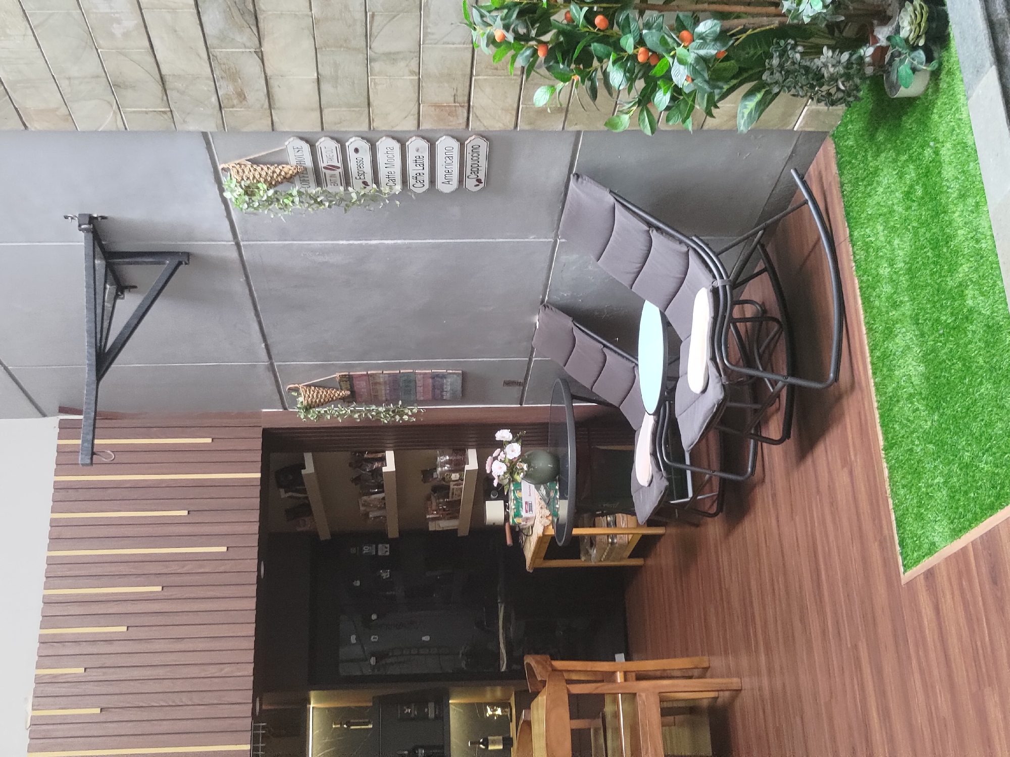 The image depicts an outdoor seating area at a coffee house in Depok, Indonesia. The scene features two dark gray upholstered lounge chairs facing a round black table. A small artificial plant with orange berries is positioned to the right of the chairs. Behind the chairs, a gray textured wall is adorned with a series of white signs indicating coffee menu items like "Espresso," "Caffe Mocha," "Caffe Latte," "Americano," and "Cappuccino." A hanging artificial green plant is situated above the signs. To the left, a wooden table and chairs are visible, suggesting an indoor or adjacent seating area. The floor is covered with dark brown wood-look laminate, and a patch of artificial green grass is laid out in the foreground. The overall ambiance is that of a casual and modern coffee shop.