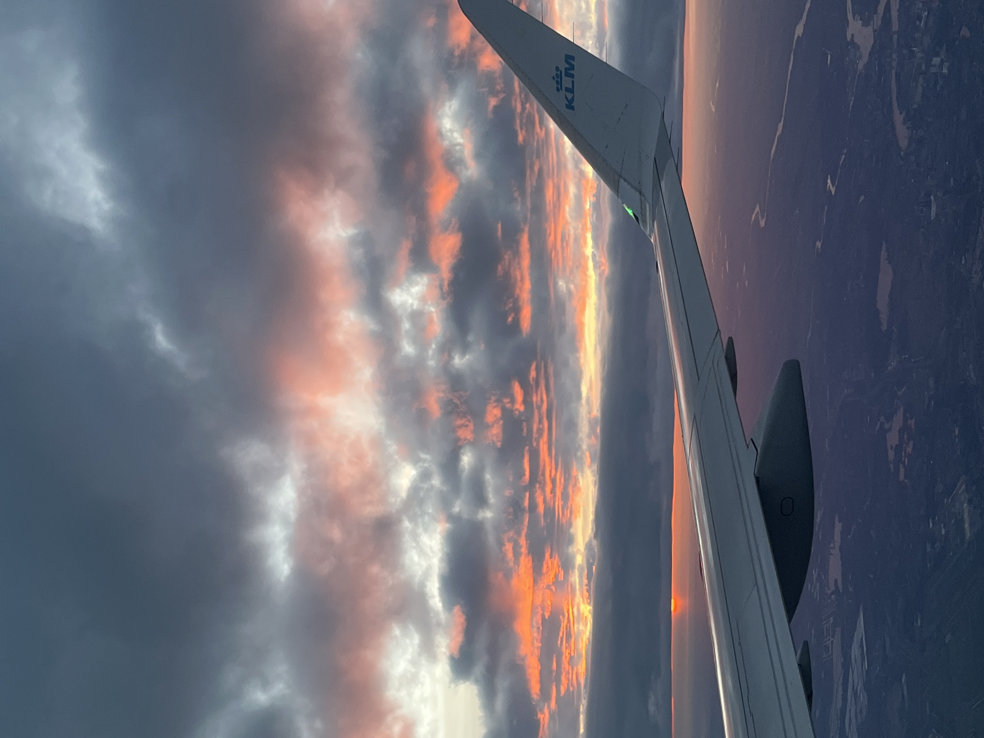The image shows a view from an airplane window, likely a KLM flight, at sunset. The wing of the plane is visible in the right side of the frame. The sunset creates a dramatic sky with a mix of dark, grey clouds and bright orange and pink hues. Below the wing, the ground is visible, appearing as a dark expanse with some lighter areas that could be water or land. The picture was not taken in Maassluis, Netherlands. It was taken from an airplane.