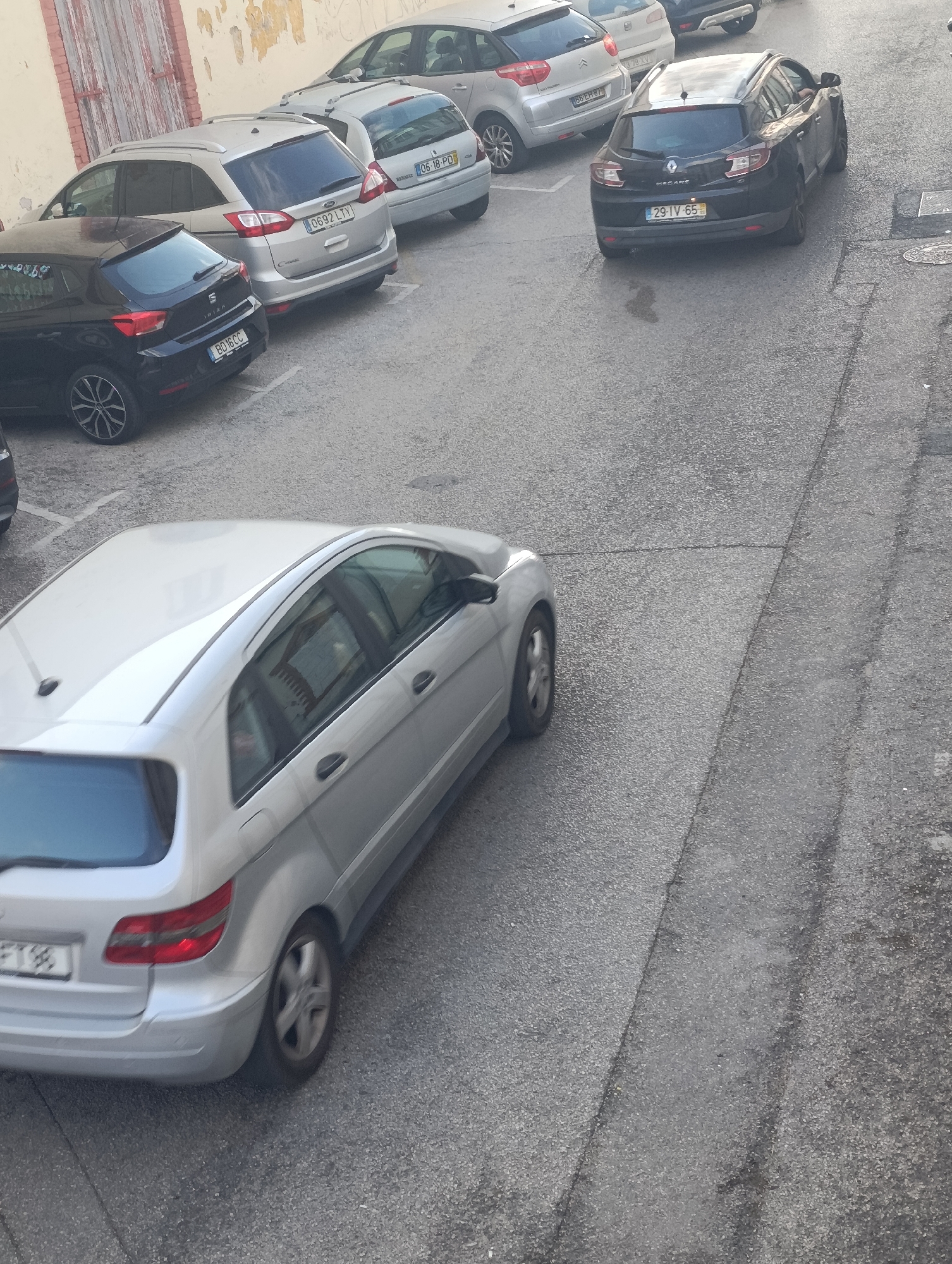 A high-angle, eye-level, medium shot captures a row of parked cars on a paved street in Setúbal, Portugal. The cars are of various makes and models, including a silver Mercedes-Benz B-Class, a silver Ford C-Max, a black SEAT Ibiza, and a black Renault Mégane. The street is lined with a weathered brick wall on the left side of the frame, which appears to be part of a building. The pavement is asphalt, showing signs of wear and tear, with visible cracks and a few dark stains, possibly from oil or water. The cars are parked in designated spots, indicated by white lines on the ground. The lighting suggests it is daytime, with some shadows cast by the cars. The overall scene depicts a typical urban parking area.