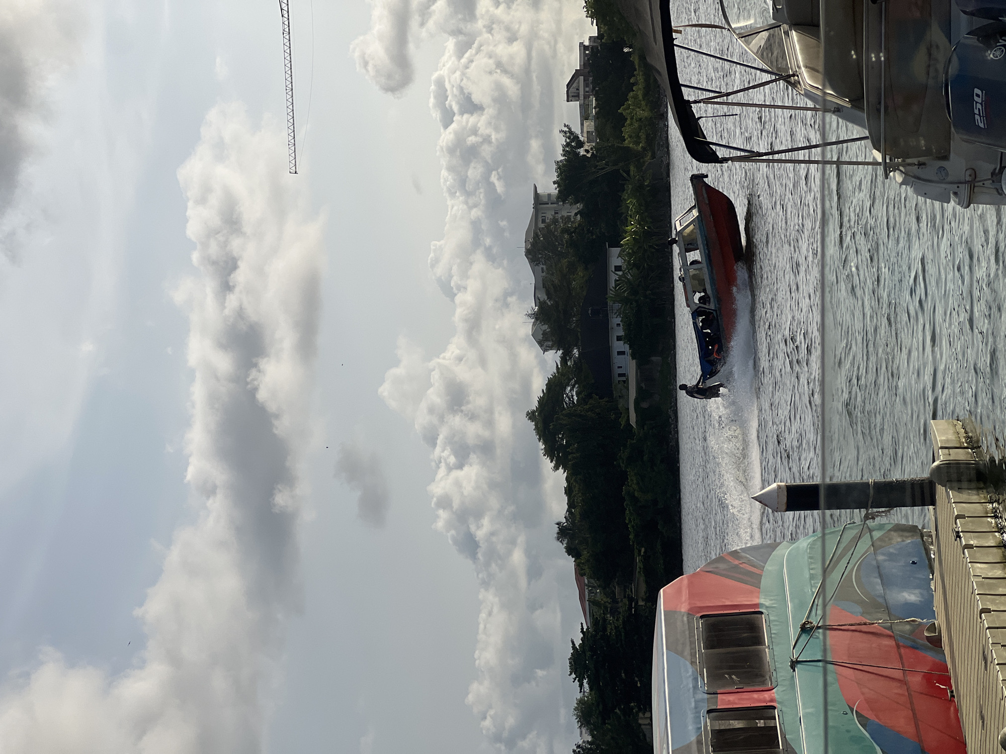 A daytime scene on the water with cloudy skies. A person is wakeboarding behind a moving orange boat. Another boat is docked alongside a pier. The scene includes buildings and trees on the shoreline. A crane structure is visible in the background. It is in Ikoyi, Nigeria.