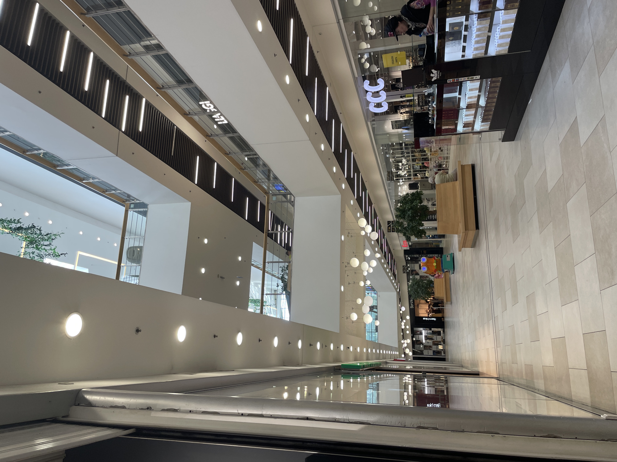 This media file captures the interior of a spacious, modern shopping mall, likely in Stara Zagora, Bulgaria, based on the provided context. The image is taken from an elevated perspective, looking down and along a multi-level structure.
The scene is dominated by clean, contemporary architecture featuring light-colored walls, white ceilings, and a light-toned tiled floor with varying shades creating a subtle pattern. On the left side of the image, a long escalator or moving walkway ascends, flanked by a white wall adorned with evenly spaced circular recessed lights. Above the escalator, the ceiling features an interesting design with dark vertical slats interspersed with linear LED strip lights, creating a sense of depth and modern illumination. A large, inverted white sign reading "ELLA 3GI" is visible near the top of this section.
On the right side, the mall opens into a wider concourse area with various shops and display areas. A prominent white sign displaying "CCC" is visible above a storefront, suggesting a shoe retailer. Further along, a glass display cabinet showcasing numerous products, possibly perfumes or cosmetics, is visible. A person in a dark top and cap is leaning over this display, appearing to browse. Below, on the main floor, several long, light-colored wooden benches are placed, with one person seated on a bench. Small potted trees are strategically placed, adding greenery to the interior landscape. Decorative spherical lights, some clustered together, hang from the ceiling in this area.
The floor is made of large, light-colored rectangular tiles. Reflections of store signs, including a reversed "BERSHKA" and a small "EXIT" sign, are visible in the glass barriers and polished surfaces on the lower level. The overall atmosphere appears bright and uncrowded, suggesting a typical shopping day.