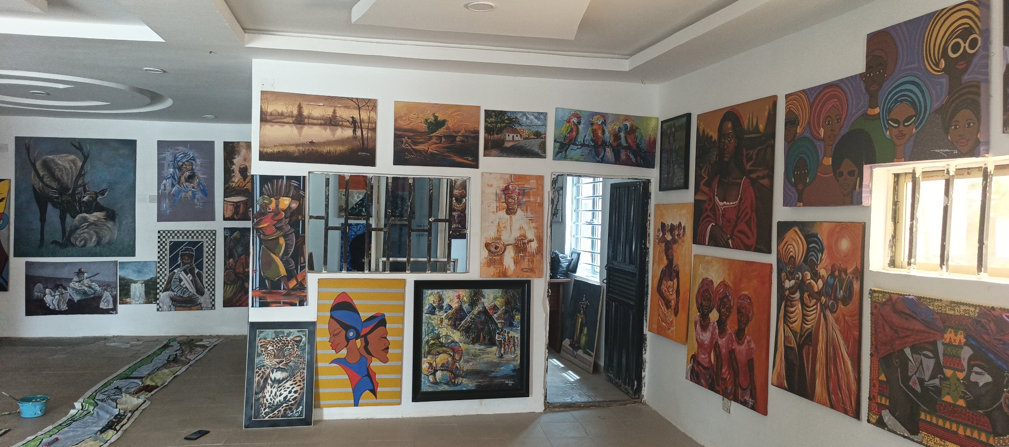An indoor view of an art gallery or studio in Akwanga, Nigeria, showcasing a dense collection of paintings on white walls. The room is brightly lit, likely by natural light from windows, with additional overhead recessed lighting. The floor is a light-colored concrete, and a long piece of fabric with painted designs, a small blue bucket, and a mobile phone lie on it, suggesting artistic work in progress.
The paintings exhibit a wide range of styles and subjects. On the left wall, prominent pieces include a large, realistic painting of a deer with a fawn, a vibrant abstract portrait of a person in blue headwear, and a painting featuring a figure playing a drum. Further down, there's a checkered pattern artwork, a scene with figures on a boat, a waterfall landscape, and a detailed portrait of a leopard.
The back wall features several landscape paintings, one depicting a serene lake with a solitary figure, and another showing a village scene. A multi-panel mirror installation with an abstract, fragmented design is centrally placed on this wall. Above a doorway, a painting of colorful birds on a branch adds a pop of color.
The right wall is adorned with numerous paintings predominantly featuring stylized African figures. Many depict women with elaborate headwraps and traditional attire, rendered in bold, warm colors like reds, oranges, yellows, and purples. Some show individual figures, while others portray groups, including what appears to be a couple. The styles range from semi-realistic portraits to more abstract and decorative compositions.
Through an open doorway on the back right, a sunlit interior with a window and a dark wooden door is visible. Another window on the far right wall lets in bright light, partially obscuring a painting. A faint, possibly incomplete, text "DO NO" can be seen reflected on the mirror. The overall impression is that of a vibrant and active artistic space.