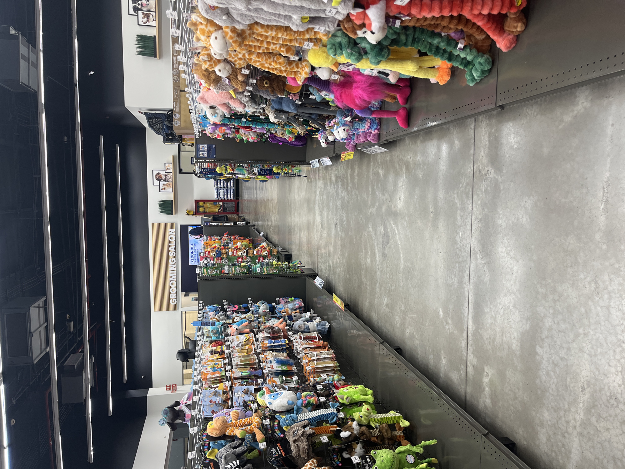 The image shows an aisle in a pet supply store. On the left, shelves are stocked with various dog toys, including plush animals and chew toys. On the right, similar shelves display a variety of plush dog toys, many of them large and brightly colored. In the background, a sign reads "GROOMING SALON." The store appears to be indoors, with a concrete floor and fluorescent lighting. There are no people clearly visible in the image. The store is located in Puebla, Mexico.
