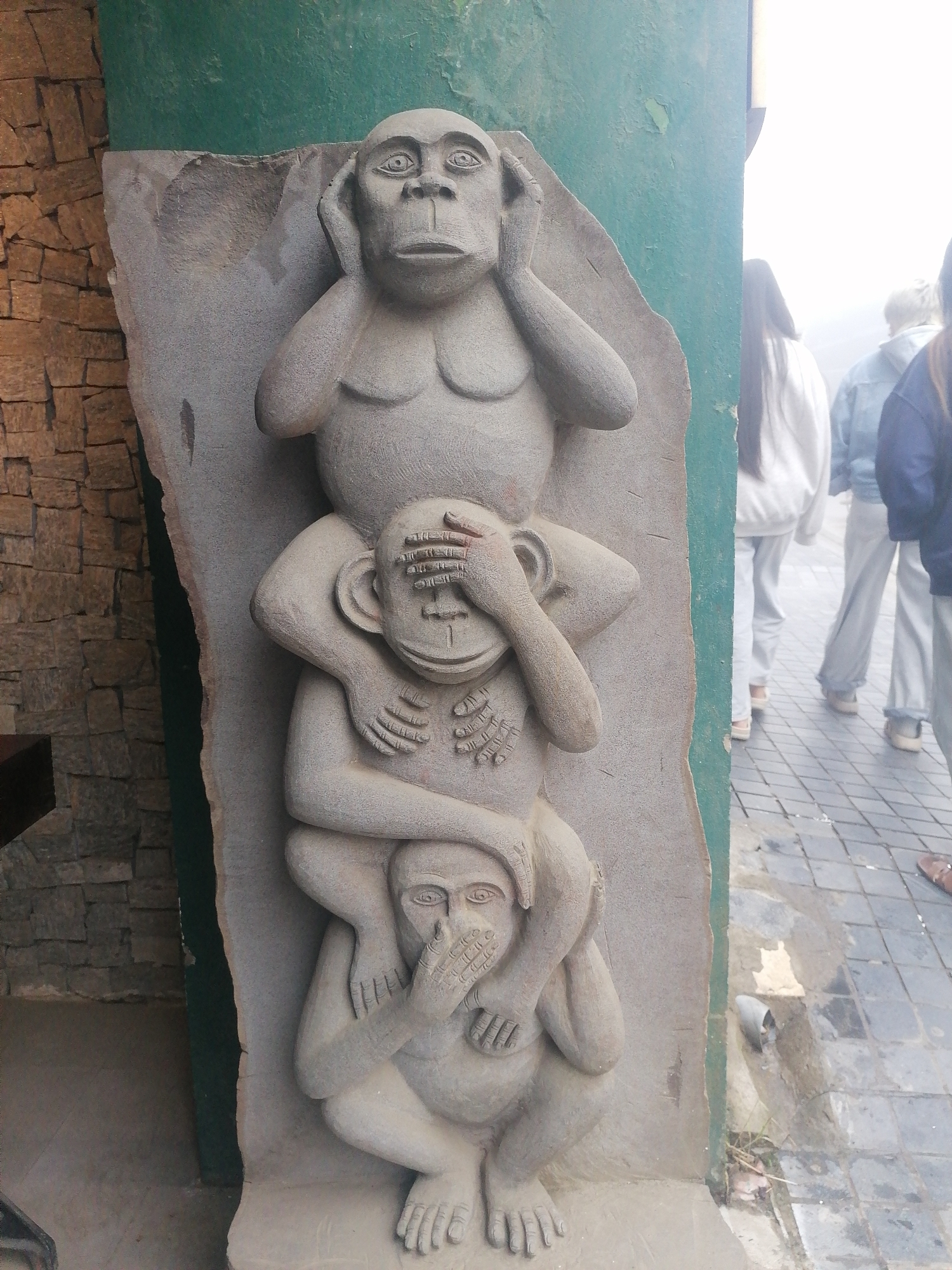 A tall stone carving, depicting the "three wise monkeys" proverb, stands vertically against a green wall on the left side of the image. The sculpture features three ape-like figures stacked one on top of the other, rendered in a natural greyish-white stone.
The uppermost figure covers its ears, representing "hear no evil." It has broad shoulders and a prominent chest, with a somber expression. The middle figure sits on the shoulders of the bottom one, covering its eyes with both hands, symbolizing "see no evil." The lowermost figure, in a squatting position, covers its mouth with its hands, embodying "speak no evil."
To the left of the statue, a wall constructed from irregular, reddish-brown stones is visible. The green wall behind the statue shows signs of wear, with some paint peeling.
On the right side of the image, beyond the green wall, several people are seen from behind, walking on a paved surface with a square tile pattern. Their figures are somewhat blurred, and the background beyond them is hazy and bright, suggesting an overcast day or atmospheric conditions often present in mountainous regions like Sa Pa. The individuals are dressed in casual attire, including hoodies and jackets. The time of day appears to be daytime, with ample ambient light. No text is visible in the image.