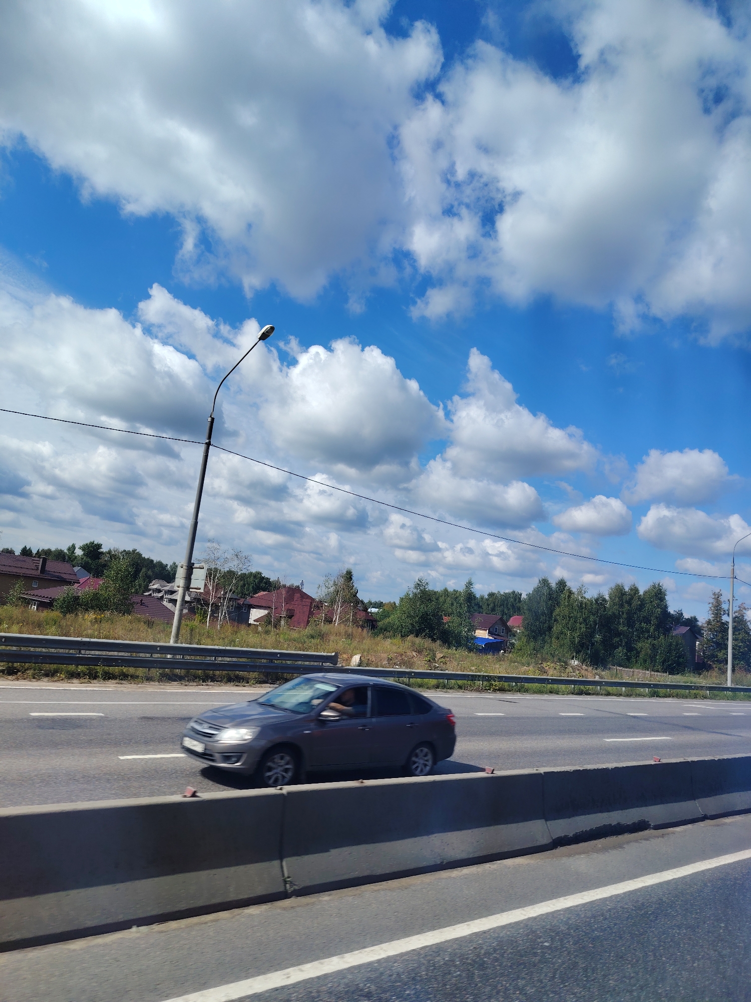 The image depicts a daytime scene on a multi-lane highway with a grey sedan in motion on the right lane. The car appears to be a Lada Granta, based on its visible features, and a person is visible in the driver's seat. The highway is bordered by a concrete barrier and a metal guardrail separating it from a grassy embankment with scattered trees and residential buildings in the background. The sky is filled with white cumulus clouds against a vibrant blue backdrop. A tall streetlamp with a curved arm is visible on the left side of the road. The setting is likely Lozhki, Russia, given the architectural style of the houses and the presence of a Lada vehicle.