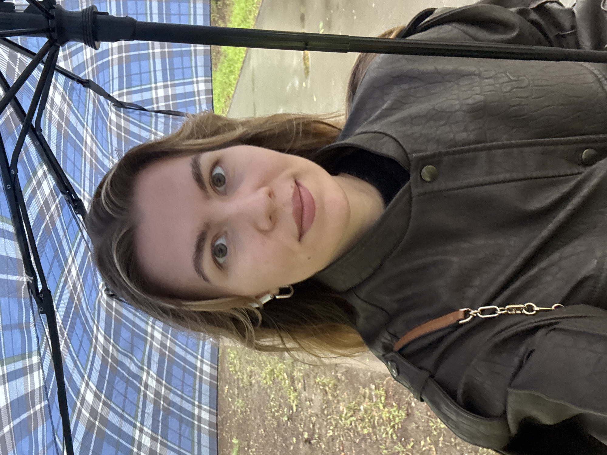 A woman is taking a selfie outdoors under a blue and white checkered umbrella. She is wearing a dark brown textured jacket with a tan strap attached to a gold chain detail on the left shoulder. She has medium-length brown hair and is wearing silver hoop earrings and white wireless earbuds. The background shows a paved path and grassy areas with some fallen leaves. The lighting suggests it may be an overcast day. The umbrella is open above her head, with its dark pole visible on the right side of the frame.