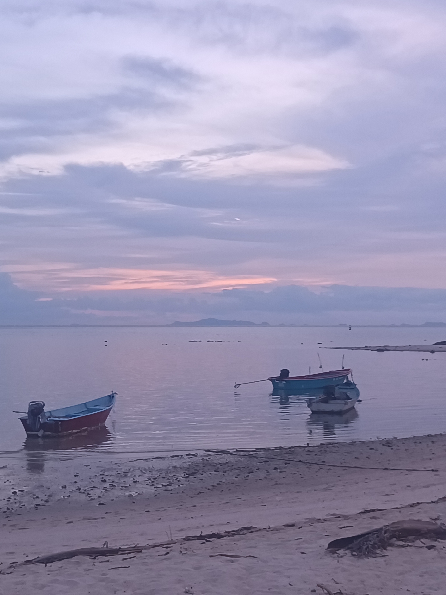 The image depicts a tranquil coastal scene in ตำบลเกาะพะงัน, Thailand, likely during either dawn or dusk. The sky is largely overcast with stratocumulus clouds, displaying a gradient of soft lavender, pale pink, and light gray hues, with the most intense pink concentrated along the horizon. A thin, crescent moon is visible in the upper left quadrant of the sky, partially obscured by clouds.
The calm, shallow water of the sea, stretching across the middle ground, reflects the muted colors of the sky, appearing as a smooth expanse of lavender-gray. Several small, dark landmasses, possibly islands or distant hills, are discernible along the horizon line. In the mid-distance, three small buoys or markers are sparsely distributed in the water.
Three small boats are present in the foreground and mid-ground. To the left, a boat with a dark red hull and light blue interior is partially beached in very shallow water, an outboard motor clearly visible at its stern. Further to the right, two additional boats float in slightly deeper water. The larger of these two has a teal-colored hull and a dark interior, featuring a long tiller or steering pole extending rearward. Adjacent to it, a smaller boat with a light gray or white hull and dark interior is also present. All boats appear to be small, local fishing or utility vessels.
The foreground consists of a sandy beach, characterized by light brown and gray tones. The sand contains numerous small pebbles and shell fragments, particularly near the waterline. A dark, linear object, possibly a rope or piece of driftwood, lies horizontally across the lower right portion of the beach. The scene is devoid of any human presence or activity.