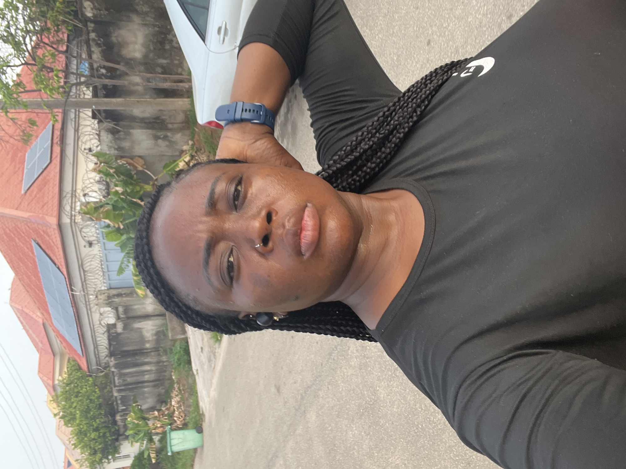 A dark-skinned woman wearing a black long-sleeved shirt and blue smartwatch is lying on the side of a street in Abuja, Nigeria. She has braided hair and a nose ring. A car and a house with solar panels on the roof are visible in the background.