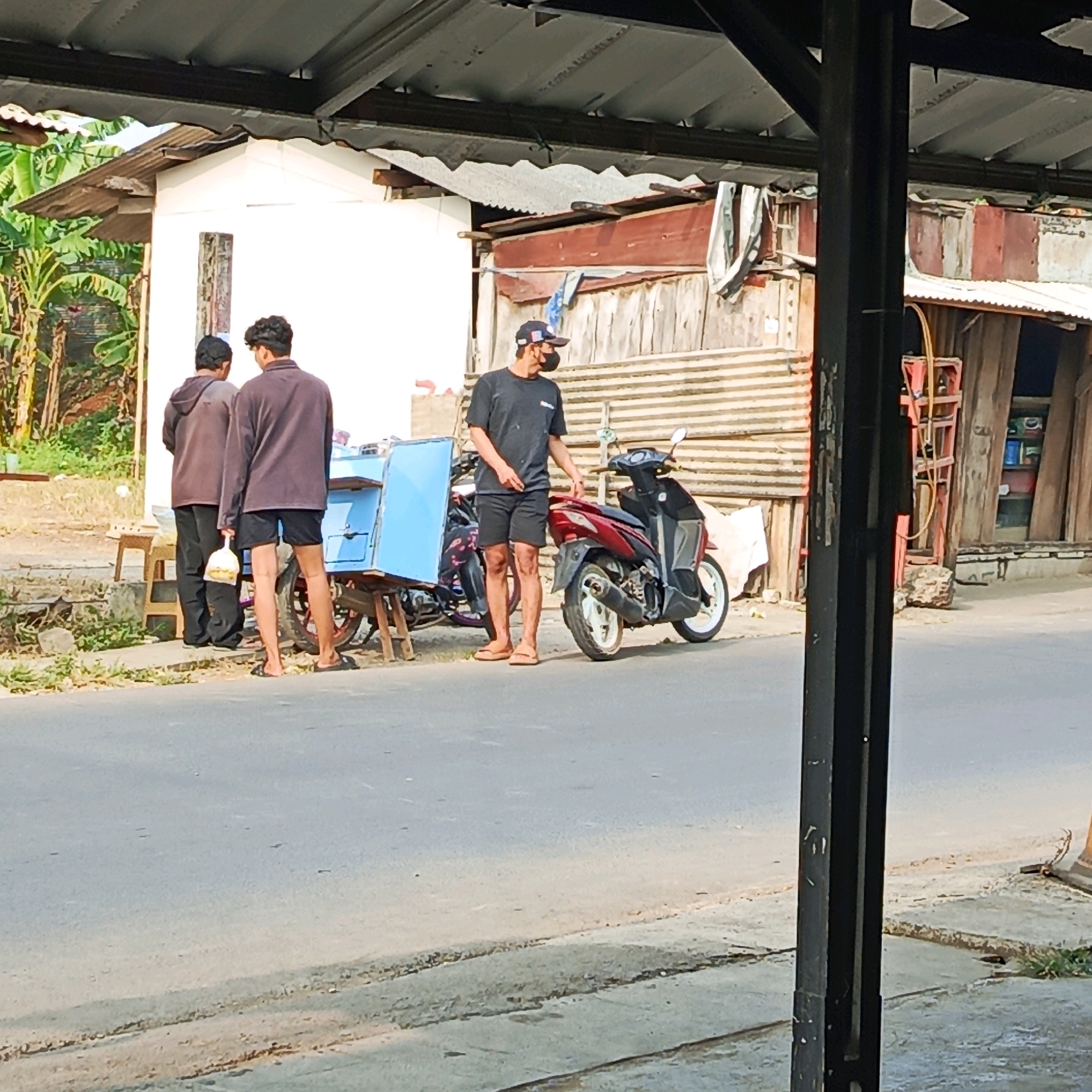 In Karawang, Indonesia, on a paved street under a corrugated metal awning, three individuals are observed. Two men stand near a light blue cart, one wearing a brown long-sleeved shirt and dark pants, and the other a similar brown shirt with black shorts, holding a white plastic bag. The third man, wearing a black t-shirt, black shorts, and a cap, stands next to a red scooter. The background features a white building with a corrugated metal roof and various wooden structures. Lush green vegetation is visible behind the buildings. The scene appears to be an outdoor marketplace or street-side vendor area.