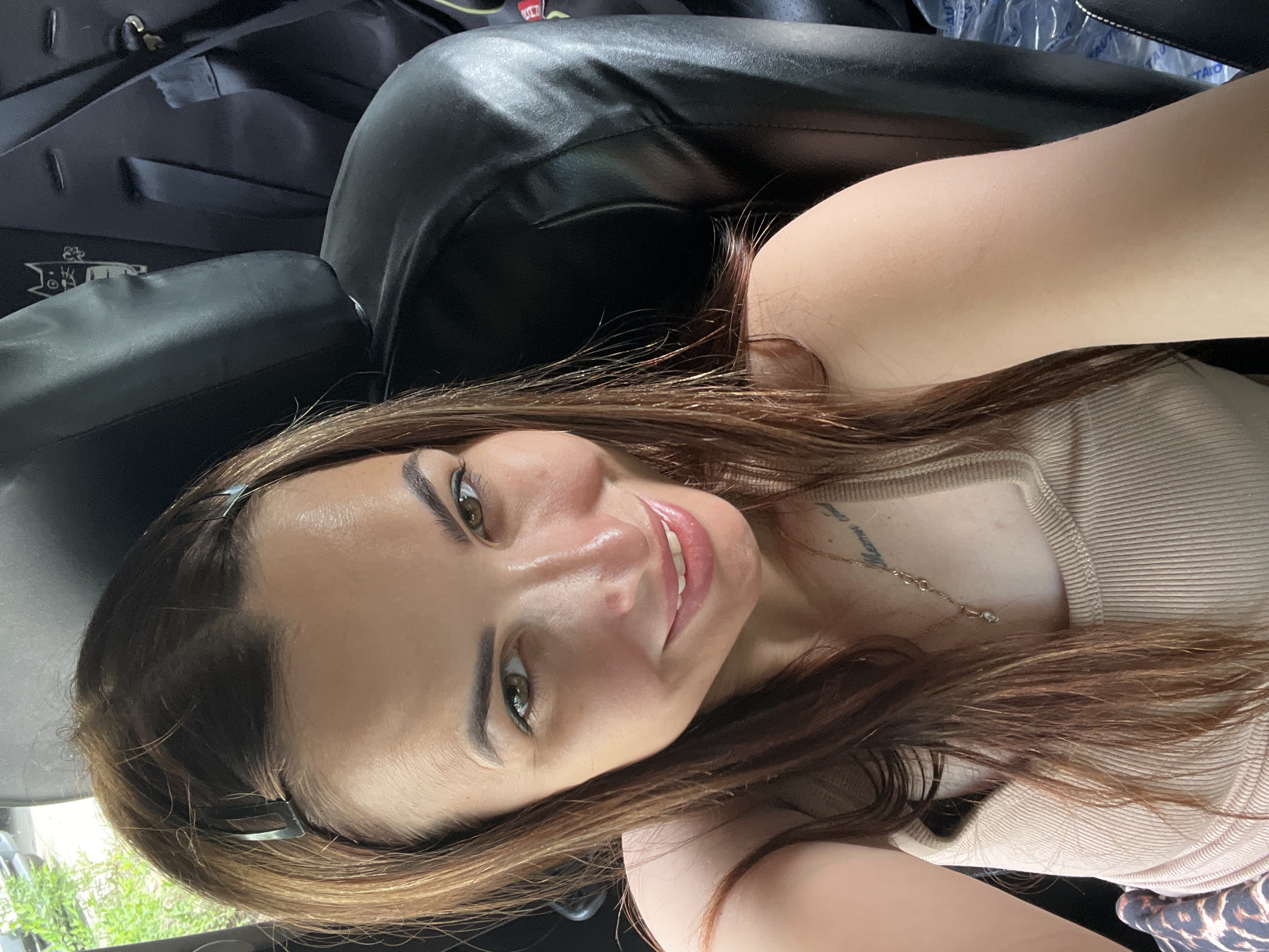 A woman with brown hair, wearing a beige tank top and a necklace, is sitting in the backseat of a vehicle, her body angled towards the camera. She has a light complexion and is smiling. Her hair is partially pulled back with clips. The background consists of black leather seats and a portion of a black garment with a white cartoon cat design. The scene appears to be outdoors, with greenery visible through a window on the left side of the image.
