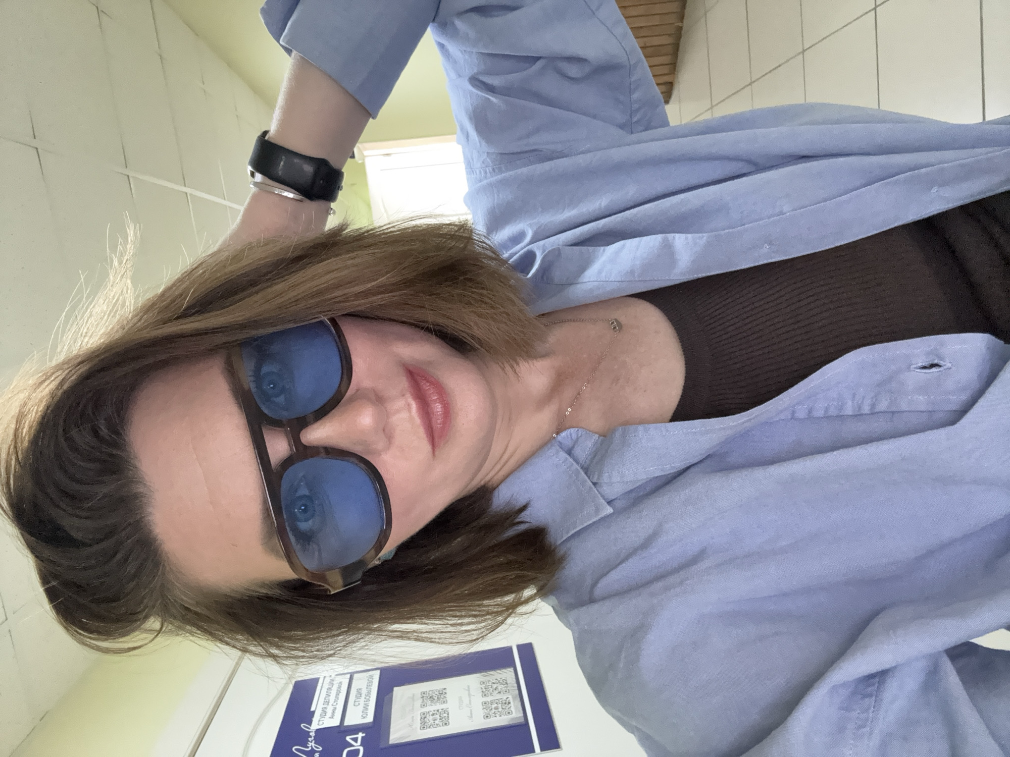 A woman is taking a selfie in what appears to be a public hallway. She has brown hair styled in a tousled bob and is wearing large, dark-rimmed glasses with blue lenses, a light blue button-up shirt open over a brown tank top, and a black watch on her left wrist. A silver necklace with a crescent moon pendant is visible around her neck. To her left, a blue poster is affixed to a wall, featuring text in Russian and QR codes. The hallway has white tiled floors and pale green walls. The background shows part of a doorway or interior space with wooden accents. The image is likely taken indoors in a building.