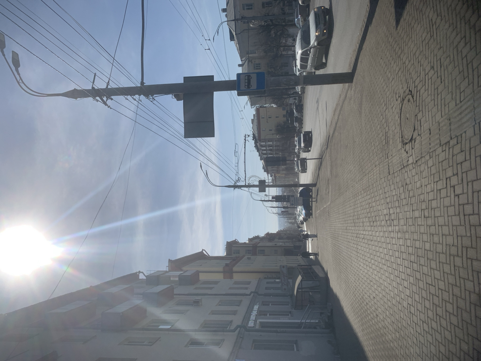 The image shows a street scene in what appears to be Izhevsk, Russia. The road is lined with buildings of varying heights and architectural styles, constructed with light-colored materials. A sidewalk paved with a light gray brick pattern runs along the right side of the street. A bus stop sign with a blue bus icon is present. Multiple cars are parked along the street, while others drive by. Power lines crisscross overhead, connected to light poles. The sun shines brightly in the sky, creating a glare. Several pedestrians are visible walking on the sidewalk.