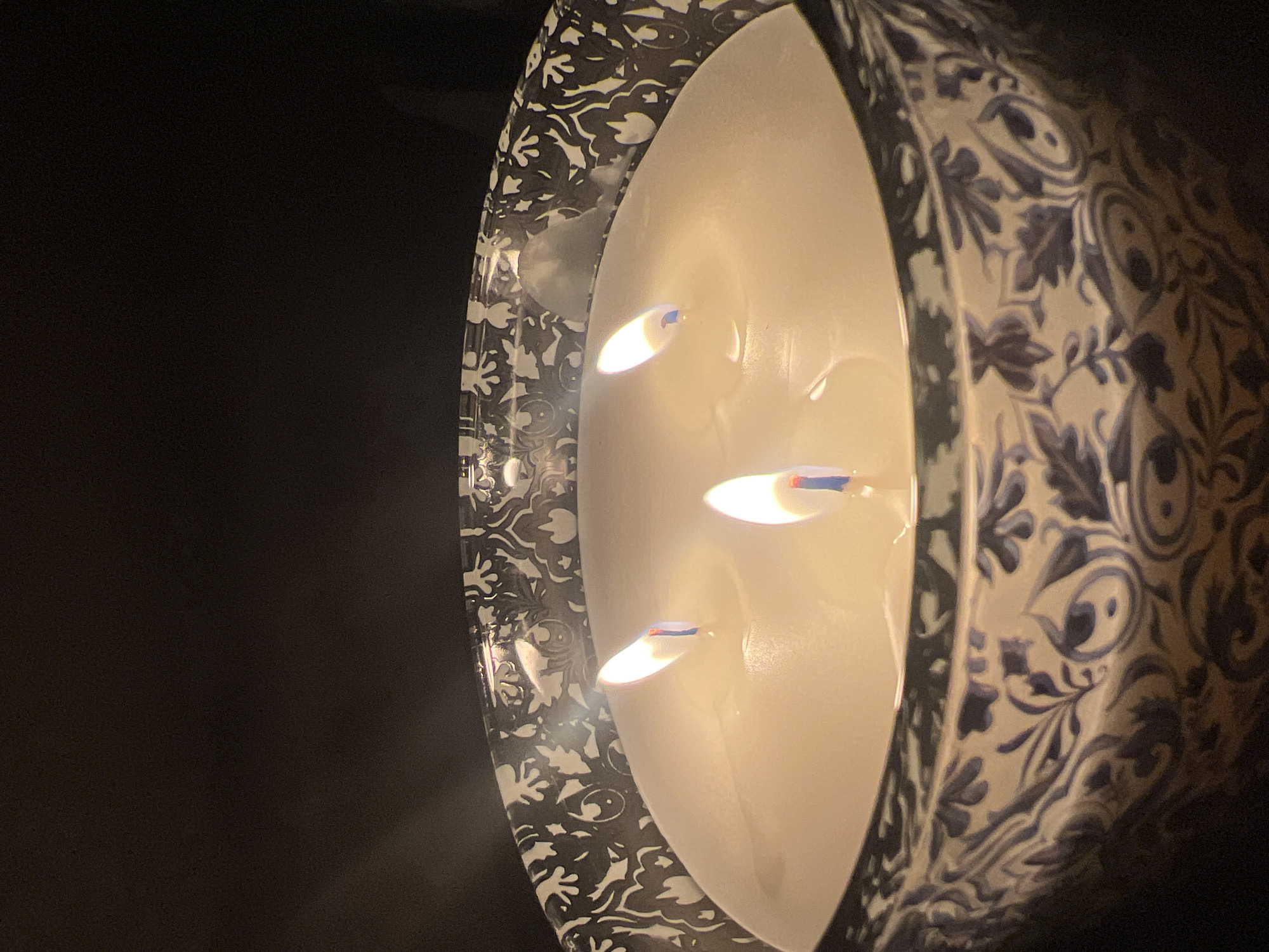 A lit, triple-wick candle sits in a patterned ceramic container. The container has a cream-colored base with a black and white floral design on one side and a blue floral pattern on the other. The candle's wicks are lit, casting a warm glow against the light-colored wax. The background is dark. No definitive signs indicate it is taken in Sofia, Bulgaria.