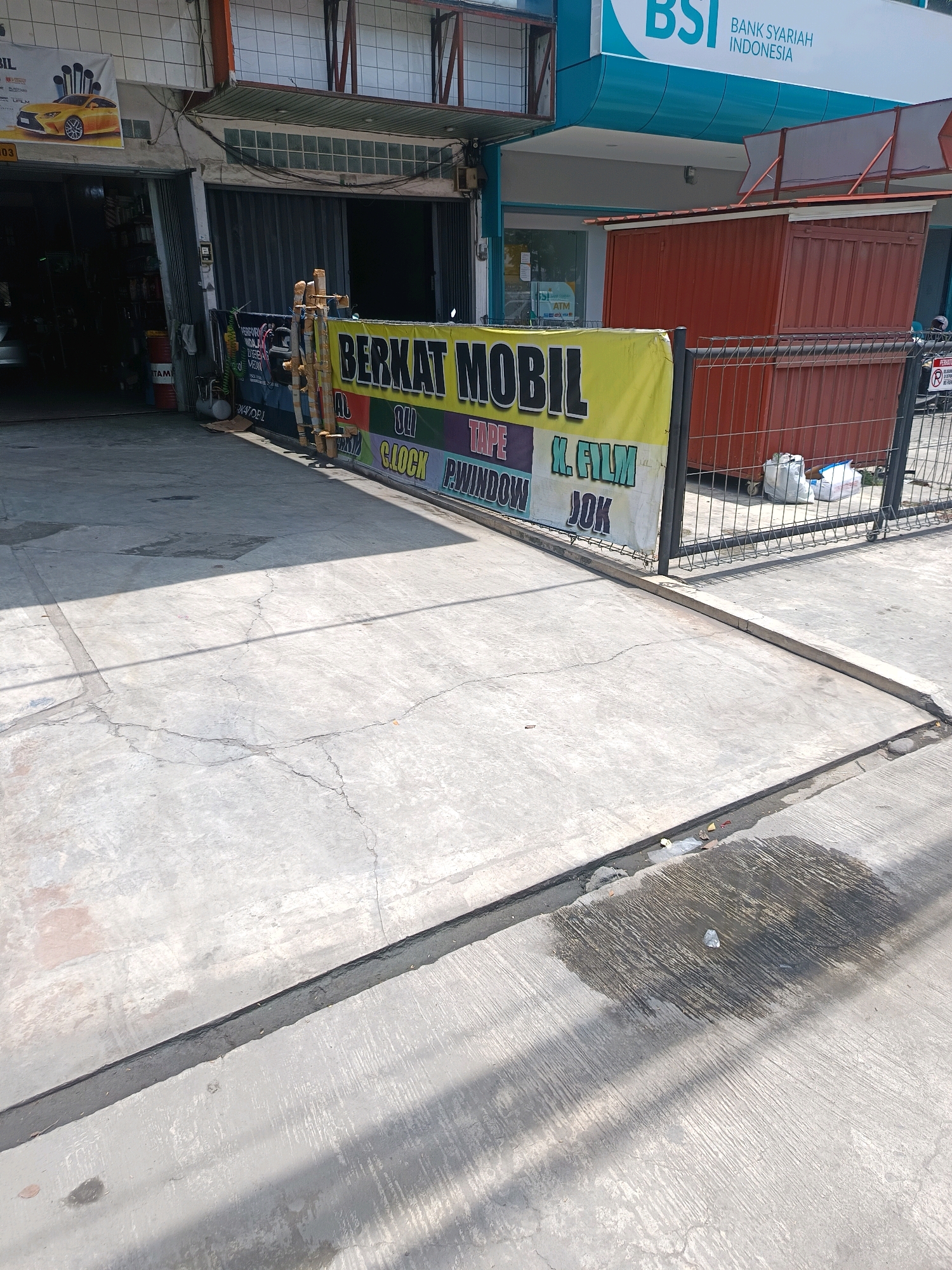 The image captures a street-level view of a commercial area in Percut Sei Tuan, Indonesia. In the foreground, a cracked concrete sidewalk and driveway are visible, with some debris and a damp patch on the ground. A black drainage channel runs along the edge of the sidewalk.
Dominating the mid-ground is a large yellow banner with black text that reads "BERKAT MOBIL," indicating a car-related business. Below this, smaller text lists services such as "OLI," "CLOUK," "TAPE," "K. FILM," "WINDOW," and "JOK." To the right of the banner, a metal fence encloses a storage area with a red corrugated metal structure. Several white trash bags are piled up behind the fence.
In the background, a building with a metallic facade and several open bays is visible, suggesting it is a workshop or garage. A yellow sports car is pictured on a poster displayed on the building's wall. Above the garage area, a section of a building with a blue and white sign that says "BSI BANK SYARIAH INDONESIA" is visible, indicating a bank on the upper floor. The scene is illuminated by bright sunlight, casting shadows across the concrete surfaces.