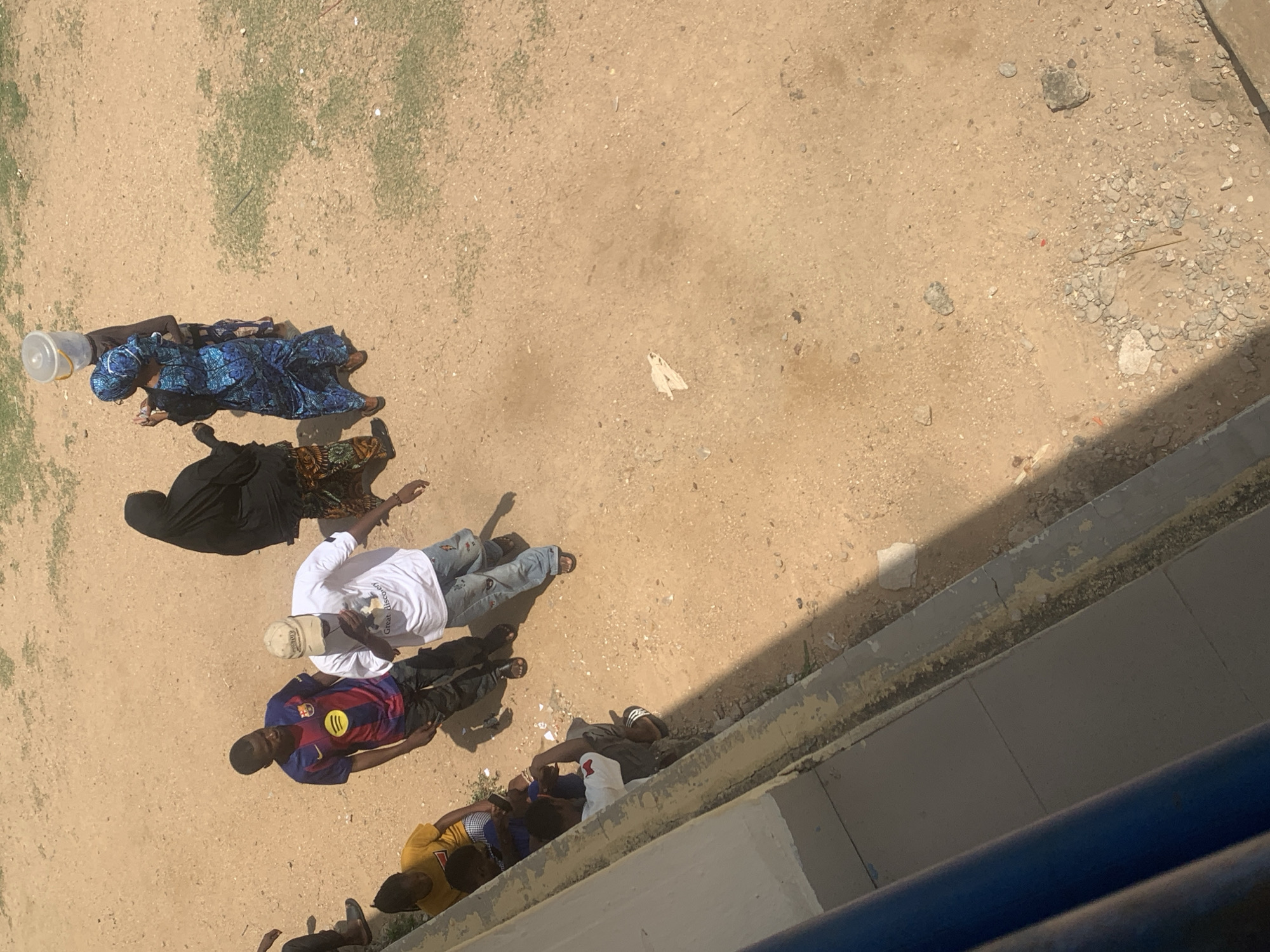 The overhead shot shows a group of people gathered on a dusty, outdoor surface. The ground is a light brown, with patches of dry green vegetation and scattered pebbles and rocks. A light-colored wall runs along the right side of the frame, casting a shadow across the ground.
In the upper left of the image, a person in a blue patterned dress and headscarf is standing next to a white bucket. Below them, another person in a black headscarf and dress is also standing. Further down, three men are walking or standing together. One man wears a white t-shirt with some visible text and blue jeans. Another man is in a blue and red striped football jersey. The third man is wearing a yellow t-shirt. Several other individuals are partially visible at the bottom of the frame, suggesting a larger group.
The activity appears to be a casual gathering or a moment of rest outdoors. The bright sunlight suggests it is daytime, likely mid-morning or afternoon given the length of the shadows. The environment appears arid and somewhat unkempt, consistent with an outdoor setting in a dry climate. The people are dressed in typical casual wear. No specific emotions are discernible from this aerial perspective. There is some visible text on the white t-shirt worn by one of the men, which appears to be an English phrase, but it is not fully legible. The scene suggests a community gathering or people in transit.