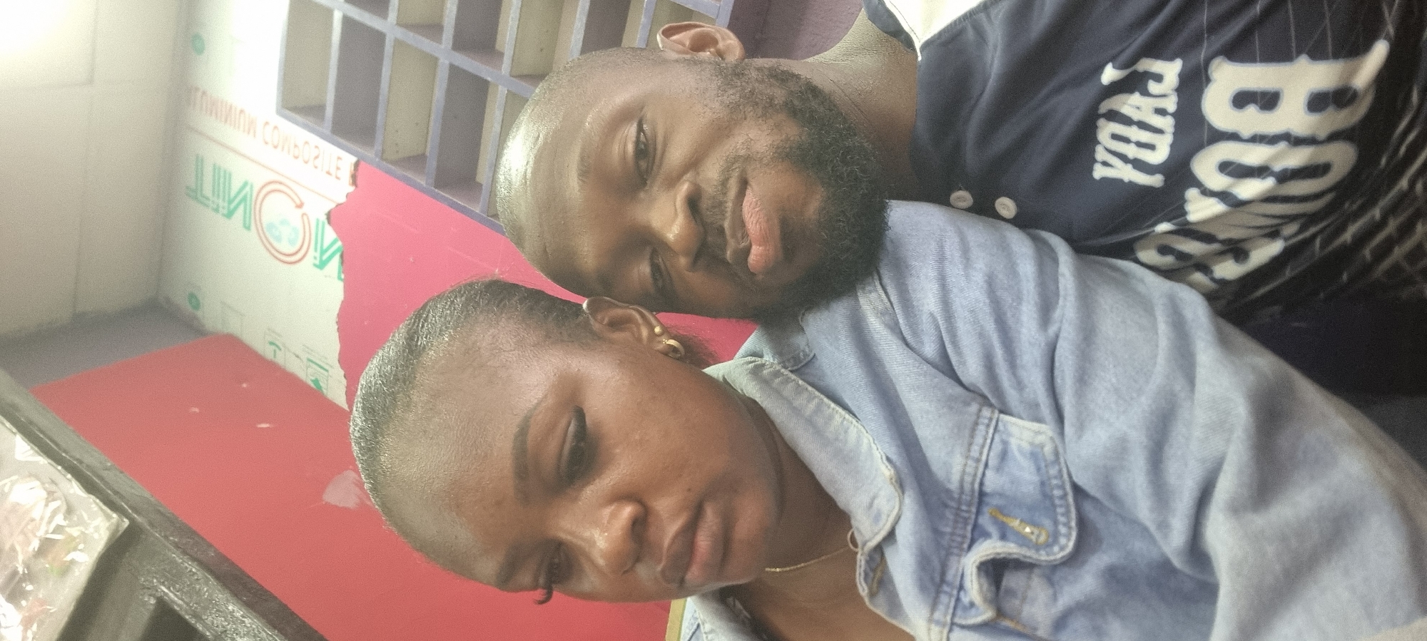 A man and a woman are posing for a selfie indoors. The man is leaning on the woman's head, and both are looking towards the camera. The woman is wearing a denim jacket. The man is wearing a dark blue baseball jersey with "BOSTON" written on it. The background shows part of a wall with a window above and red paint below. The location is presumed to be Lagos, Nigeria.