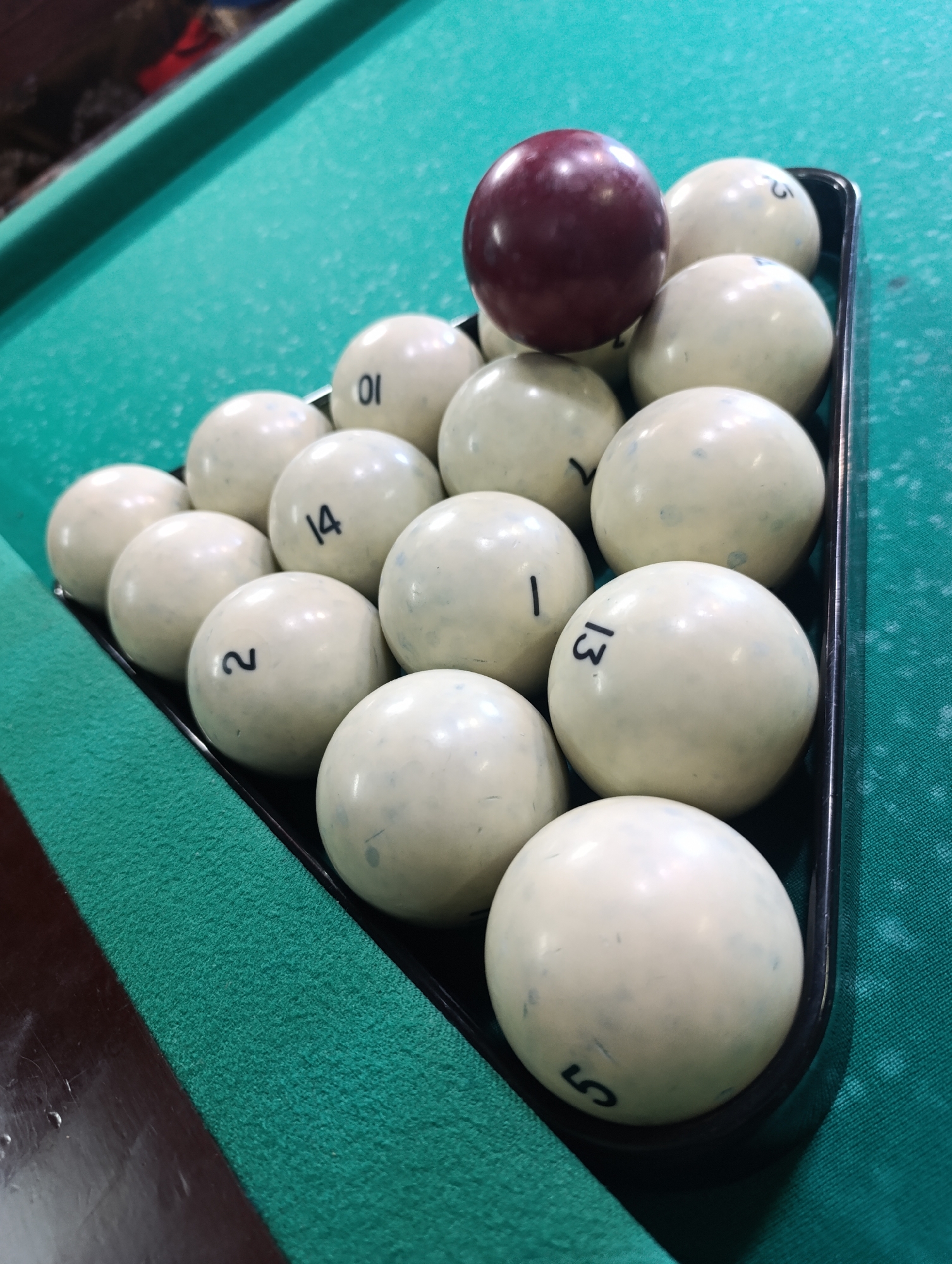 The image shows a set of billiards balls racked on a green billiards table. The balls are numbered and in a triangular rack, with a burgundy-colored ball on top. The rack sits on the green felt surface of the billiards table. This image was taken in the city of Chaikovsky, Russia.
