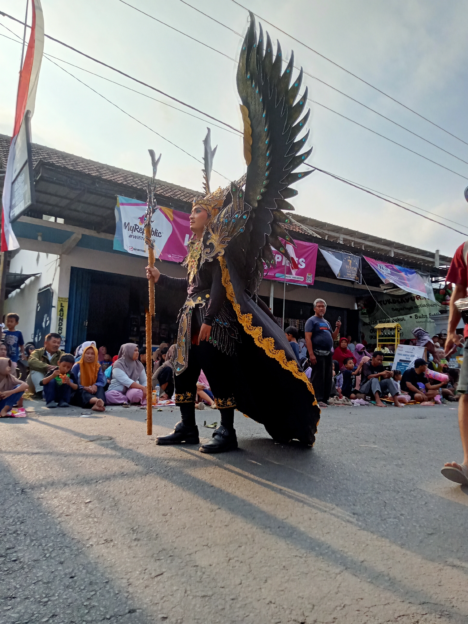 An Indonesian youth, adorned in a black and gold elaborate costume with large feathered wings and a golden headdress, stands in the middle of a street in Simo, Indonesia. He holds a wooden staff topped with decorative elements. The youth is the central figure, with a crowd of onlookers seated on the ground to his left and right. The crowd appears to be watching a parade or performance. Banners and awnings are visible on the buildings lining the street, suggesting a commercial or residential area. The sky is bright and overcast, with power lines strung across the frame. The scene is bathed in warm sunlight, casting long shadows on the asphalt road.