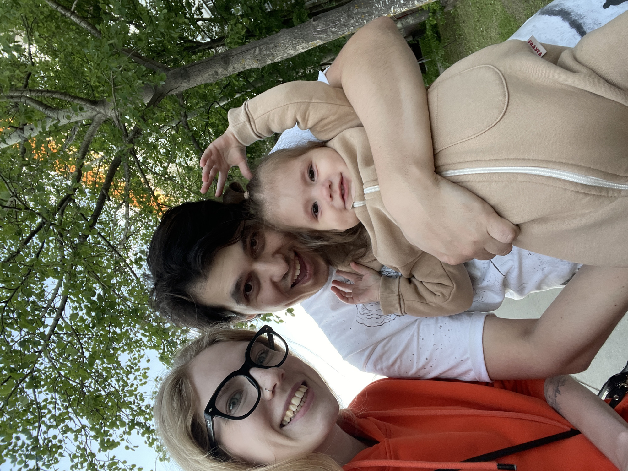 An outdoor, low-angle photograph captures a happy family of three under a canopy of green trees. In the foreground, a blonde woman with black-rimmed glasses and an orange hooded sweatshirt smiles broadly, looking directly at the camera. Faint white text "rag&" is visible on the temple of her glasses. Behind her, a man with dark hair also smiles, holding a young child. He wears a white t-shirt with a subtle pattern.
The child, likely a toddler, is dressed in a beige full-body suit (onesie) with a white zipper. A red tag or patch with white Cyrillic text "РАДУЖКА" (RADUZHKA) is visible on the child's suit. The child has hair tied in two small ponytails and looks towards the camera with a curious and happy expression, one arm extended upwards.
The background is filled with the lush green foliage of mature trees, with branches crisscrossing the frame. Patches of bright sky are visible through the leaves. In some areas, a warm, golden light filters through the canopy, suggesting the golden hour of late afternoon or early evening. The overall atmosphere conveys joy and a pleasant outdoor moment, likely in a park or natural setting within Izhevsk, Russia. The perspective suggests the photo was taken from below, possibly a selfie or an upward shot from a reclining position.