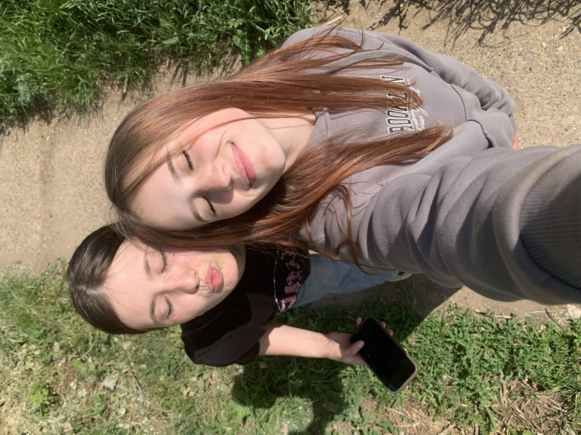 Two young women are taking a selfie in an outdoor environment. The woman on the top is wearing a grey sweatshirt with the word "BROOKLYN" visible. Her hair is long and reddish-brown. The woman on the bottom is wearing a black top and holding a black smartphone. She has dark brown hair and is making a kissing face. They are positioned close together, possibly on a path or grassy area. Green grass and patches of bare earth surround them, indicating a natural or park-like setting. The location in the city of Фоки, Russia, cannot be confirmed from the image alone.
