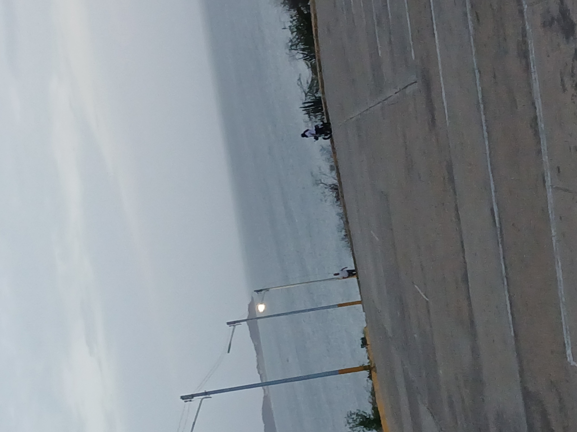 This image depicts a waterfront area in Juan Griego, Venezuela. The scene includes a paved parking lot with painted parking lines along the right side of the image. Parallel to the parking lot is a body of water, possibly the Caribbean Sea, with a visible shoreline in the distance. Streetlights line the edge of the parking lot along the water. Two people are visible near the water's edge, one standing and another one on a motorcycle. The sky is overcast and the lighting suggests it's either early morning or late evening. Vegetation grows along the shoreline dividing the water from the parking area.