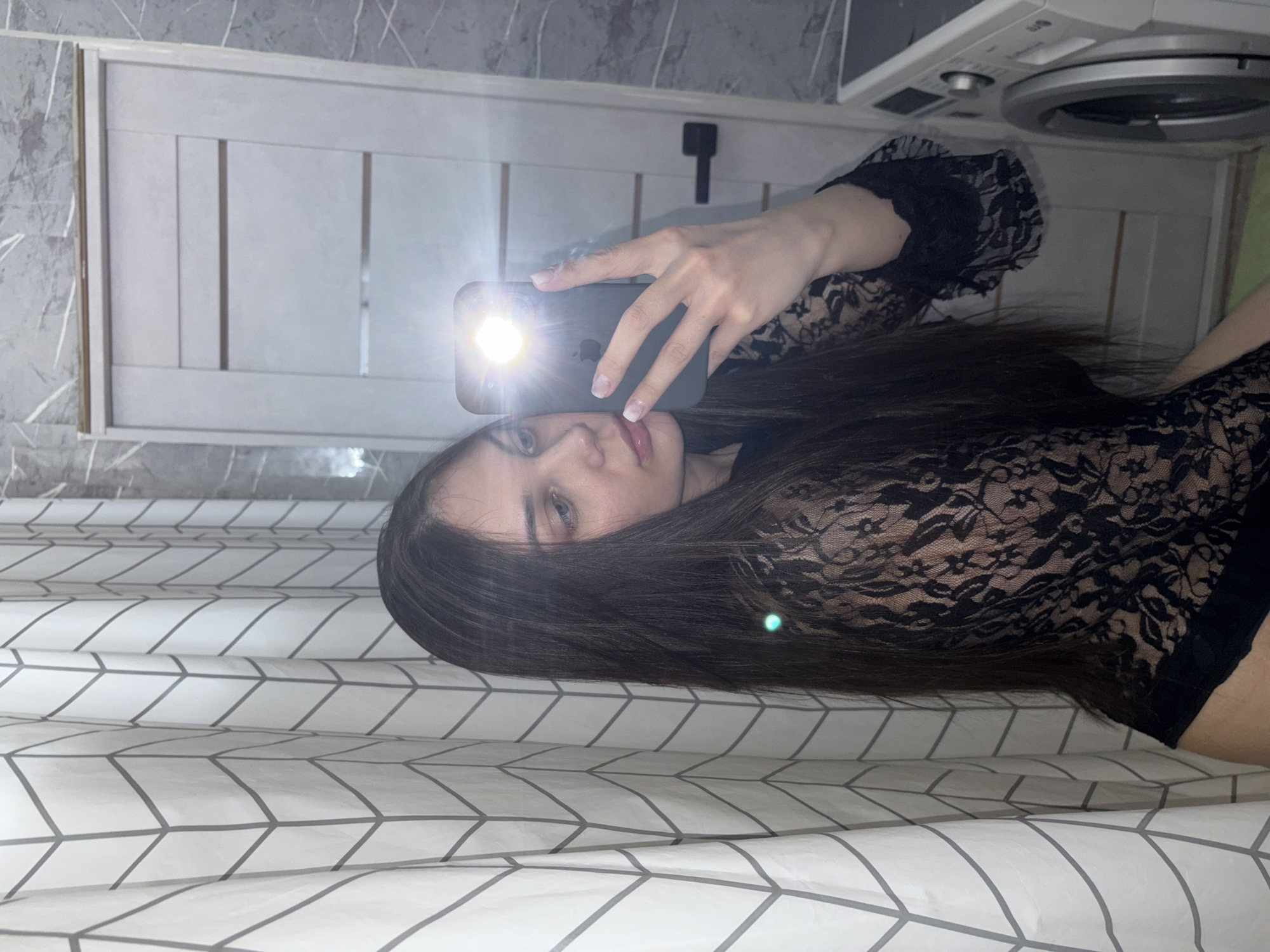 A woman is taking a selfie with a smartphone in a bathroom. She is wearing a black lace top with long sleeves. The bathroom has a white door with three recessed panels, grey marble-patterned wallpaper, and a white washing machine. The location is Chaikovsky, Russia. A shower curtain with a geometric pattern is partially visible. The phone's flash is on.