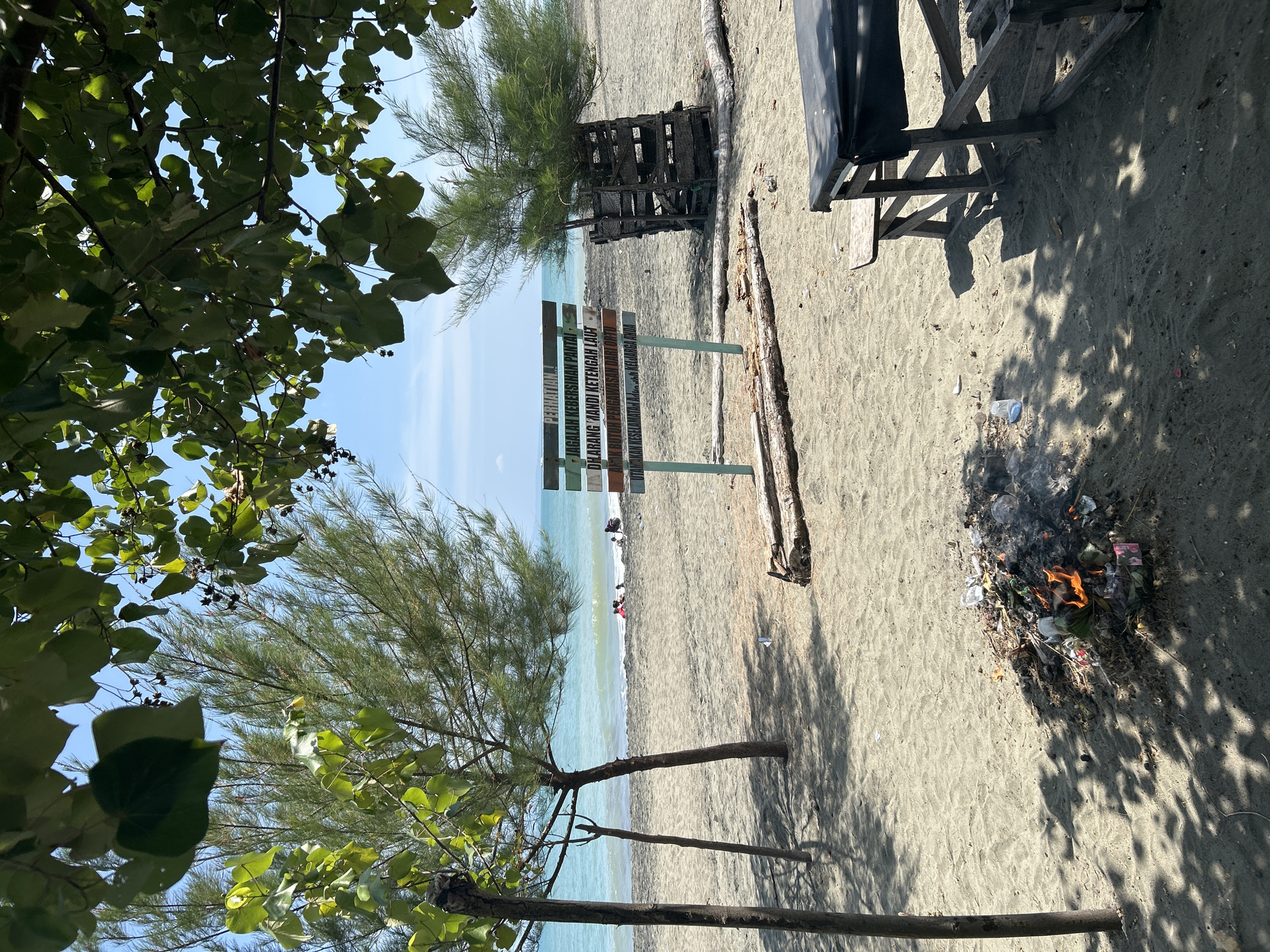 The image depicts a beach scene in Banda Aceh, Indonesia, under a clear blue sky. In the foreground, a pile of burning trash is visible on the sandy beach, with smoke rising. To the left, two thin trees with sparse green foliage stand casting shadows on the sand. In the midground, a sign with text in Indonesian reads "PERHATIAN" (Attention) followed by instructions related to maintaining beach cleanliness, prohibitions on swimming in the middle of the sea, and a reminder for parental supervision. Several small trees with dense, needle-like leaves are planted behind the sign, with one enclosed by a wooden structure. Further back, the turquoise ocean meets the sandy shore, with a few people wading in the shallow water. A simple wooden table with a black tarp covering is situated on the right side of the frame, also casting shadows. The overall environment suggests a public beach area.