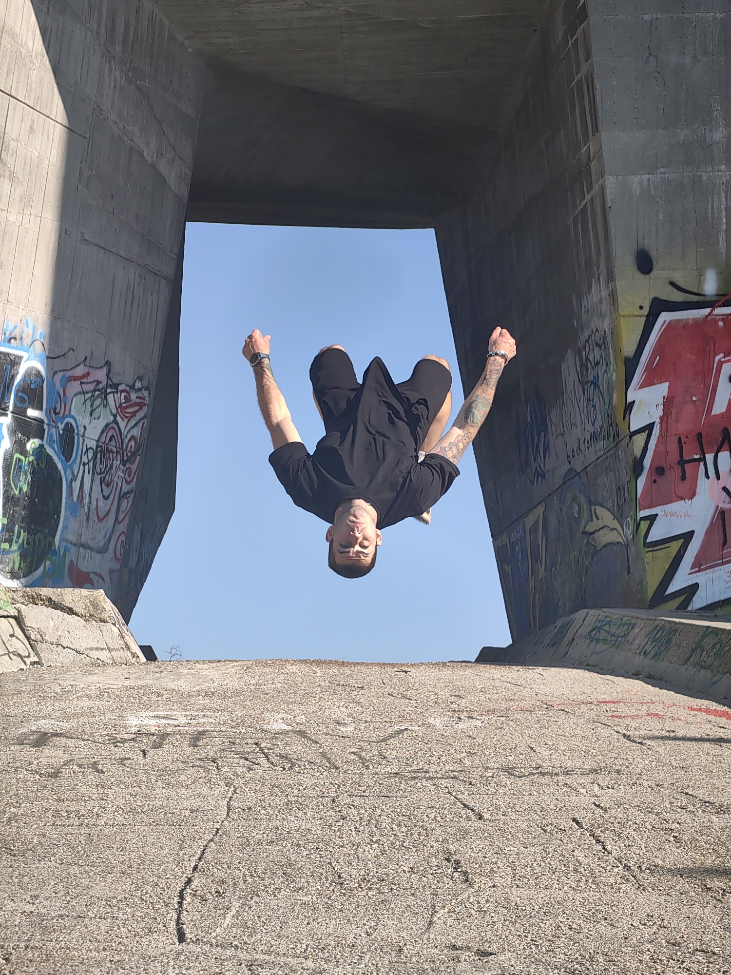 A man is performing a backflip beneath a concrete structure in Varna, Bulgaria. The man is wearing a black t-shirt and black shorts. He has tattoos on both arms and is wearing a wrist watch on his left wrist and a bracelet on his right. The concrete structure has graffiti on its sides. The background is a clear, light blue sky. The ground is concrete with cracks and worn markings. The location appears to be near the Asparuhovo Bridge.